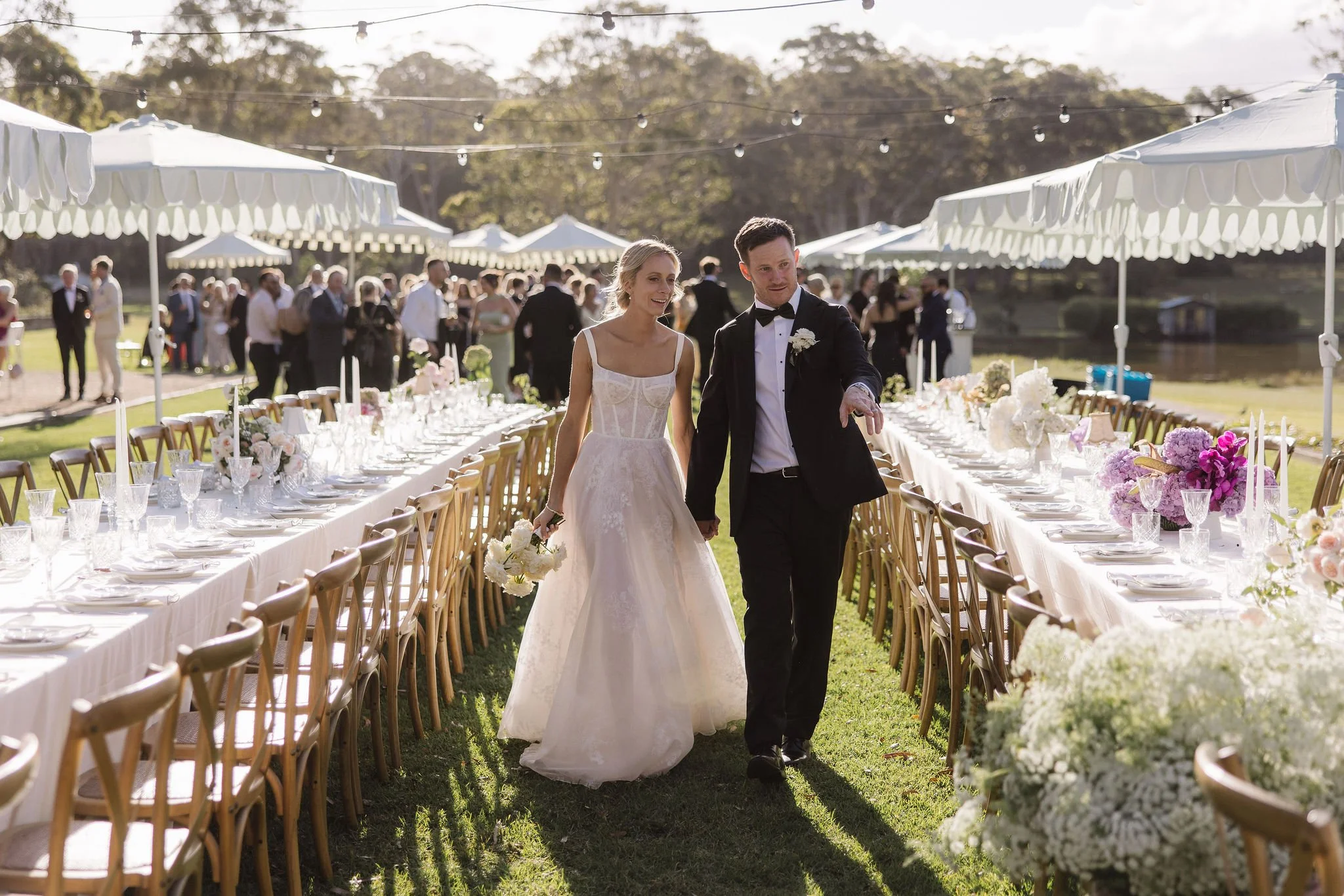 Bride and groom walking through a decorated outdoor wedding reception with long tables, white tablecloths, and floral centerpieces, under string lights with guests in the background.