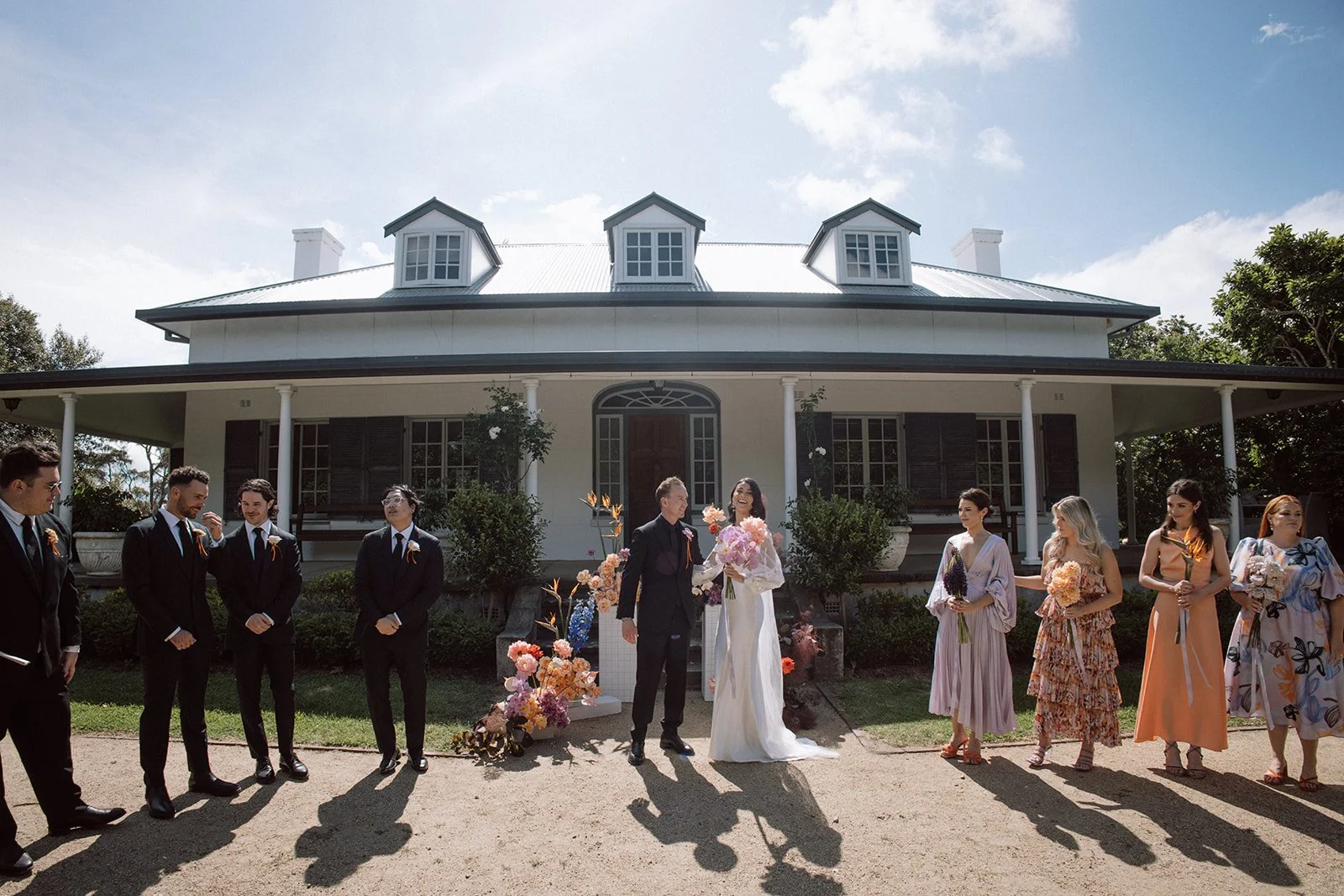 A wedding ceremony outside a large white house with multiple dormer windows. The bride and groom are standing in the center, smiling and holding hands, surrounded by friends and family. The bride is wearing a white dress and holding a bouquet of pink flowers, while the groom is dressed in a black suit. Guests are dressed in formal and semi-formal attire, holding bouquets of flowers, and smiling.
