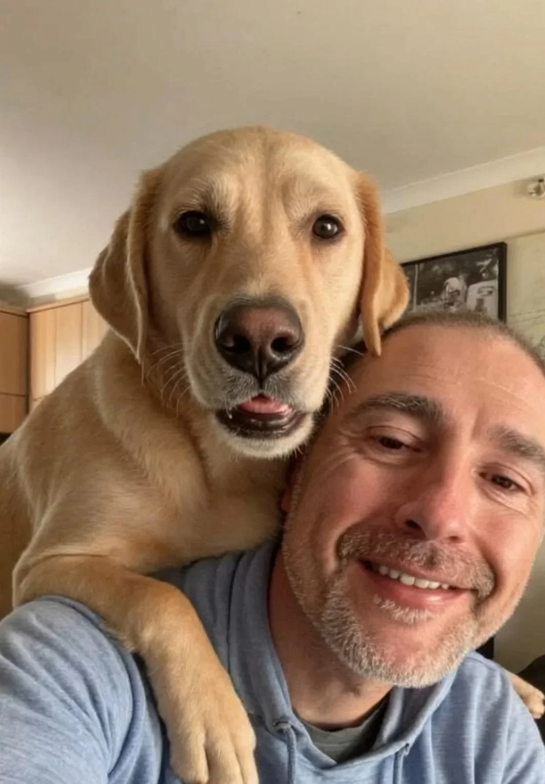 A man smiling with a yellow Labrador Retriever on his shoulder indoors.