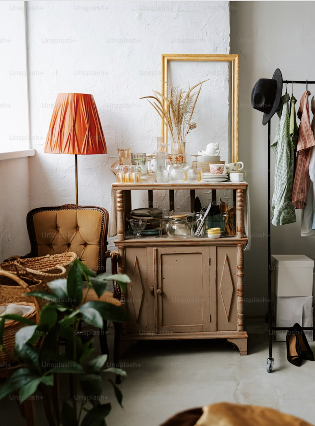 An interior scene featuring a vintage wooden cabinet with dishes and glassware, a tufted armchair with a woven basket, a tall orange lampshade, a mirror, and a clothing rack with hanging clothes and hats.
