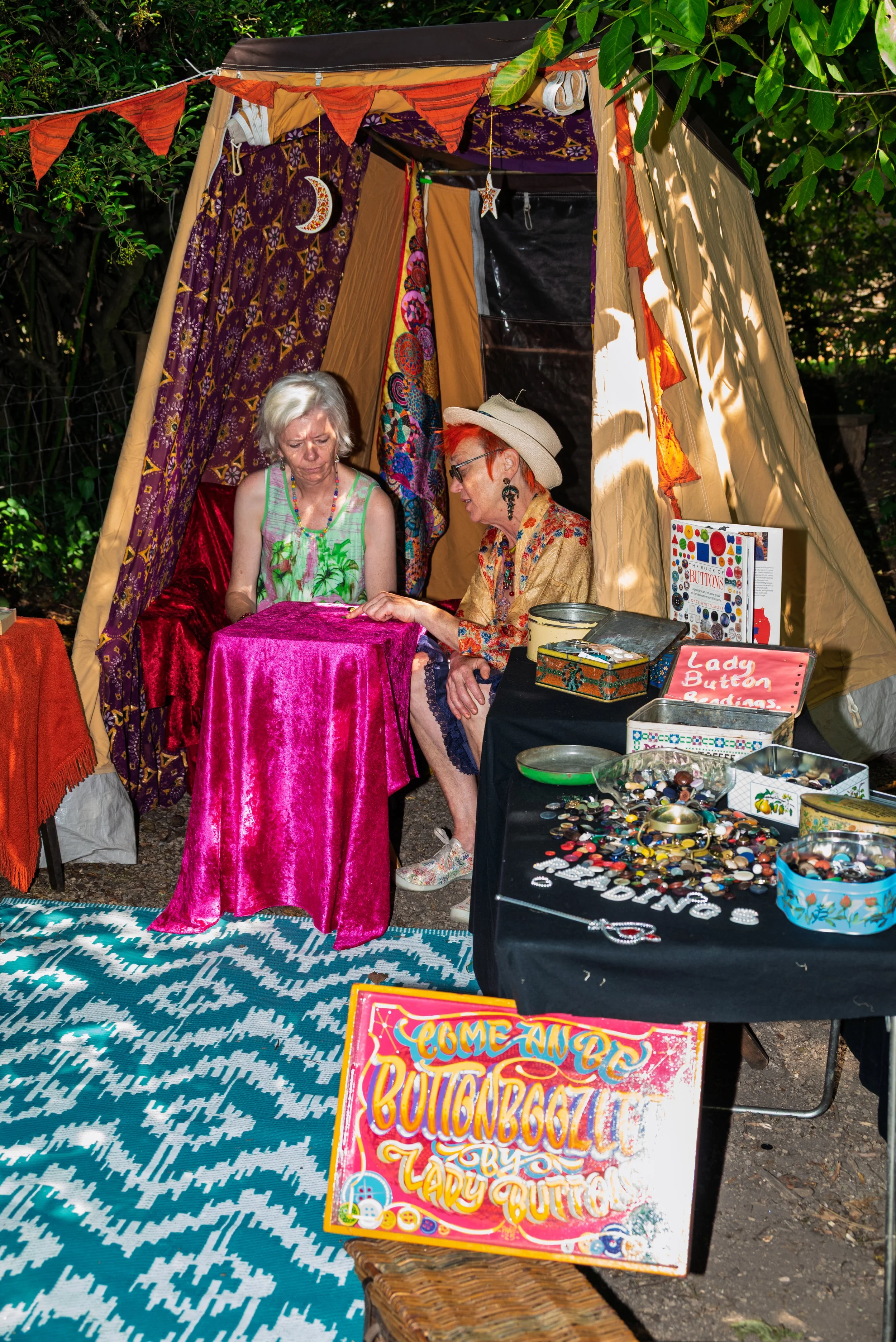 Two women sitting inside a decorative tent at an outdoor craft fair, selling button jewelry and badges, with colorful signage and a patterned rug on the ground.