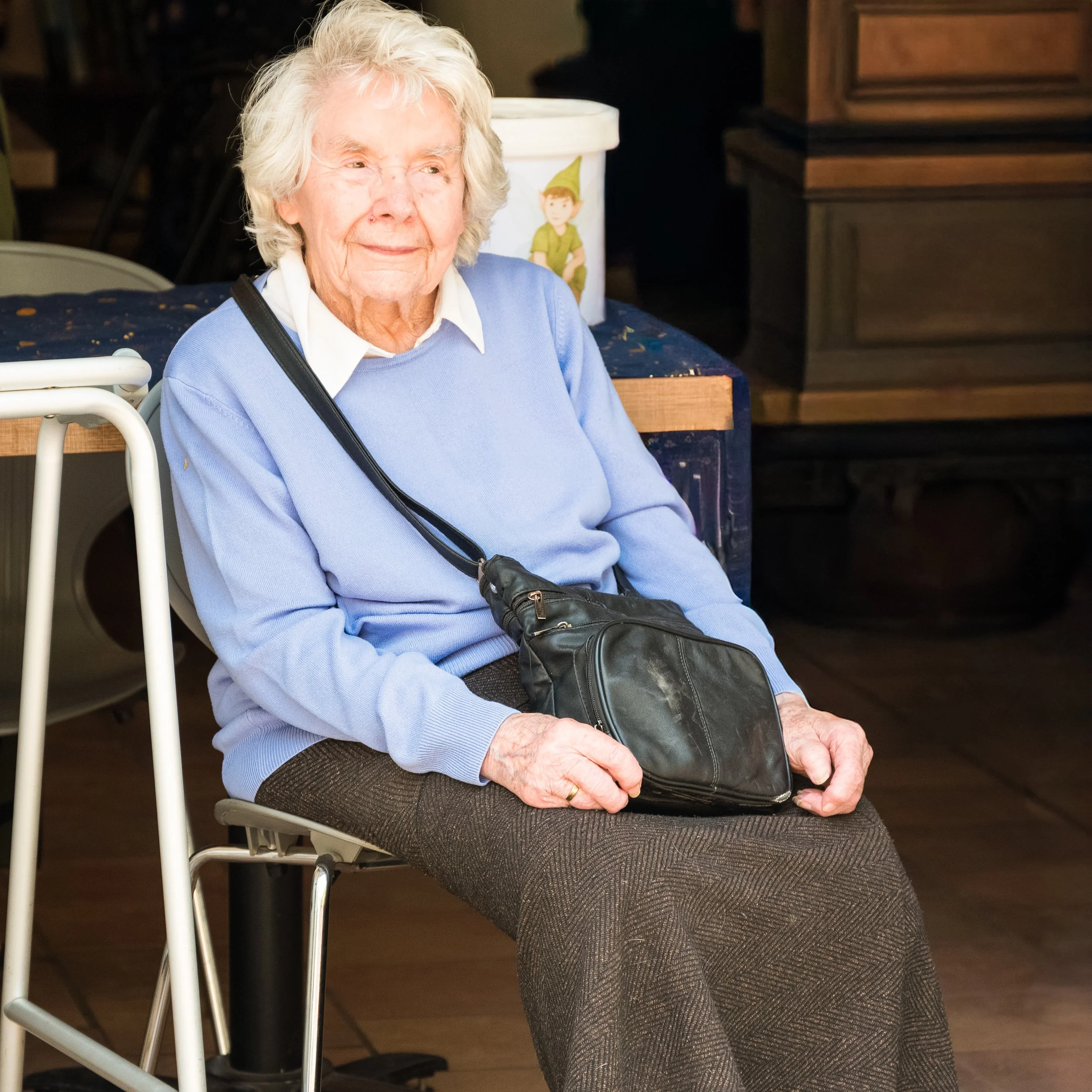 An elderly woman with white hair, wearing a light blue sweater and a white collared shirt underneath, is sitting on a chair. She has a black crossbody bag resting on her lap. In the background, there is a table with a white container featuring a cart