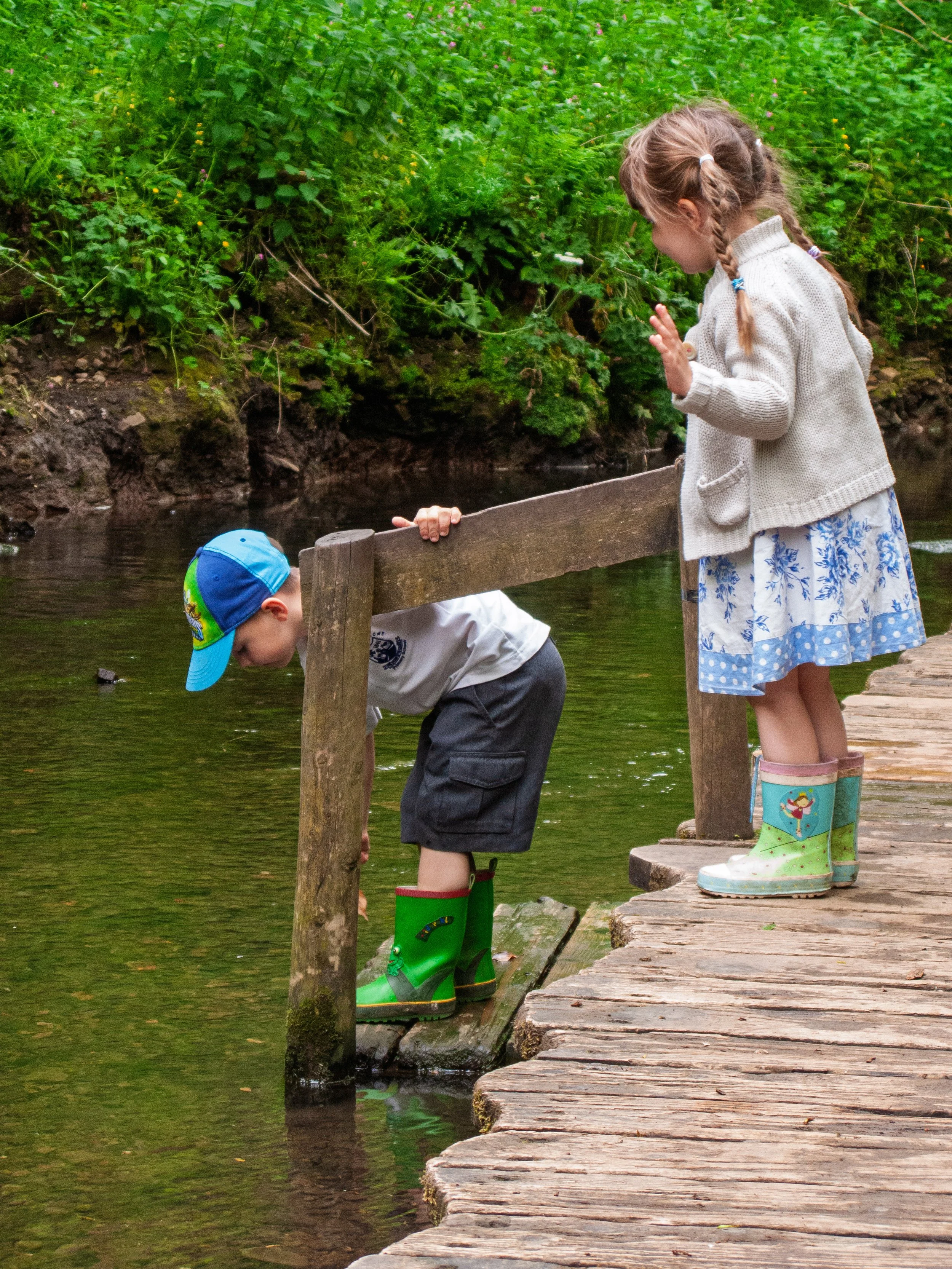 A young boy and girl by a creek, with the boy leaning over the water and the girl standing on a wooden dock, gesturing with her hand. Both children are wearing rubber boots, and the background is filled with green foliage.