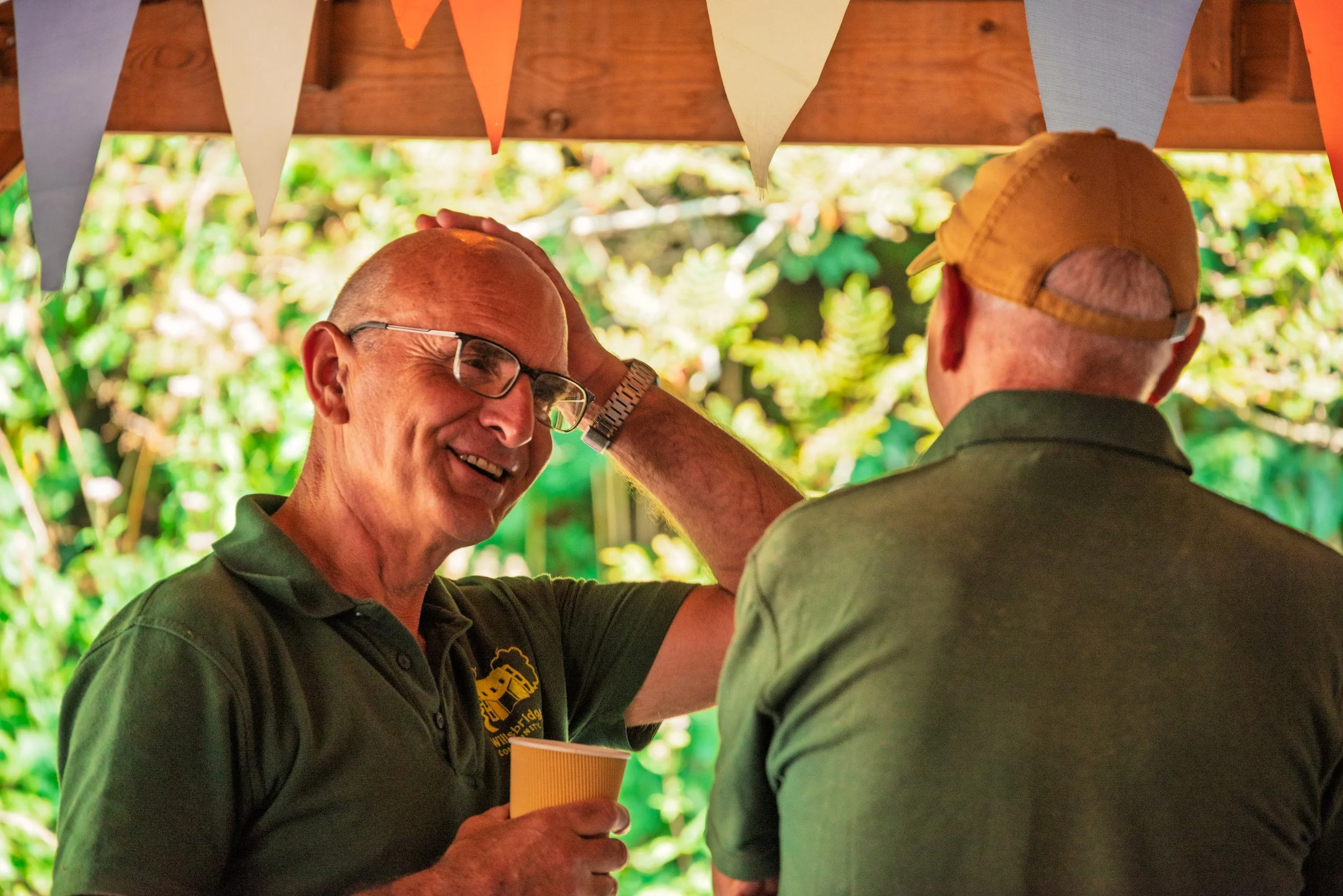 Two men at an outdoor gathering, one smiling and touching the other's head, with colorful bunting overhead and greenery in the background.