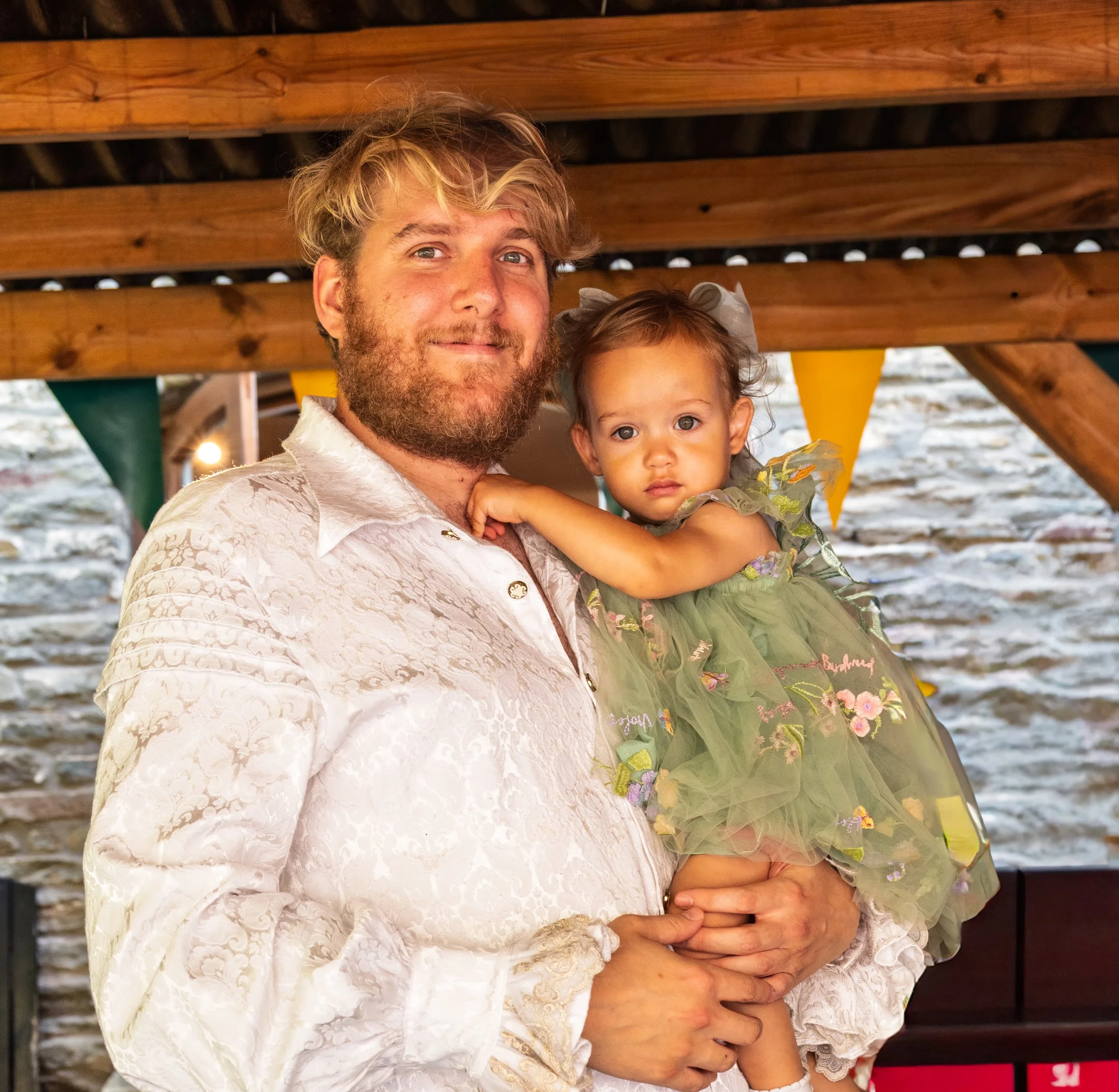 A man with blonde, curly hair and a beard holding a young girl with brown hair in a green dress. They are standing under a wooden structure with bunting in the background.