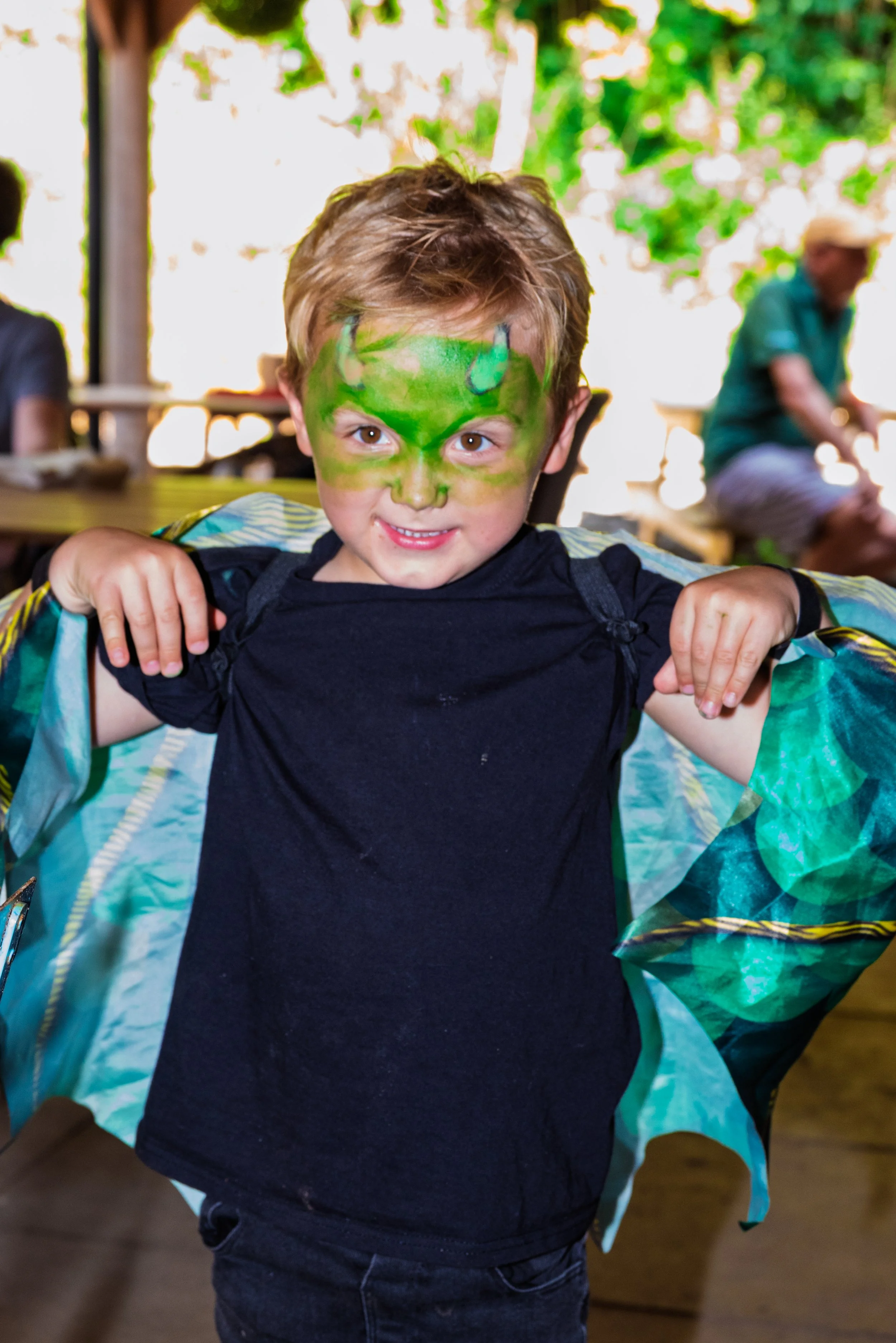 Child with green face paint dressed as a dragon, wearing a black shirt and a cape, smiling while standing indoors with people in the background.