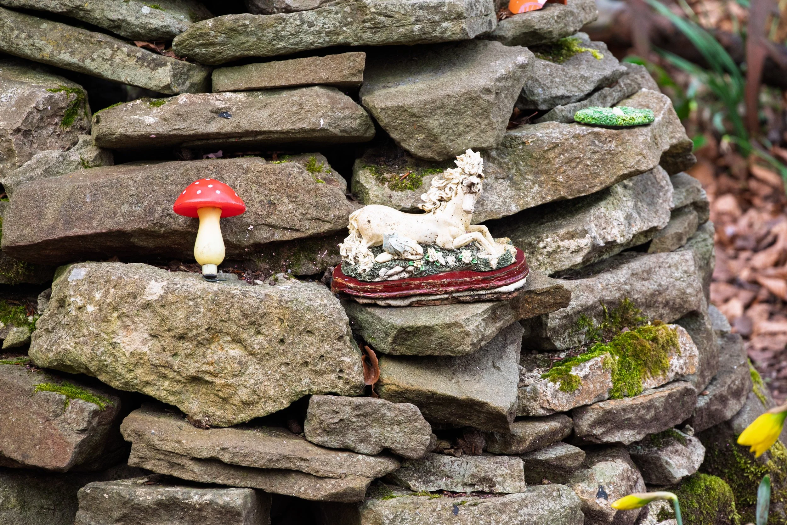 Decorative garden display with a small red and white mushroom sculpture, a white horse figurine, and a small green object on a stone wall.