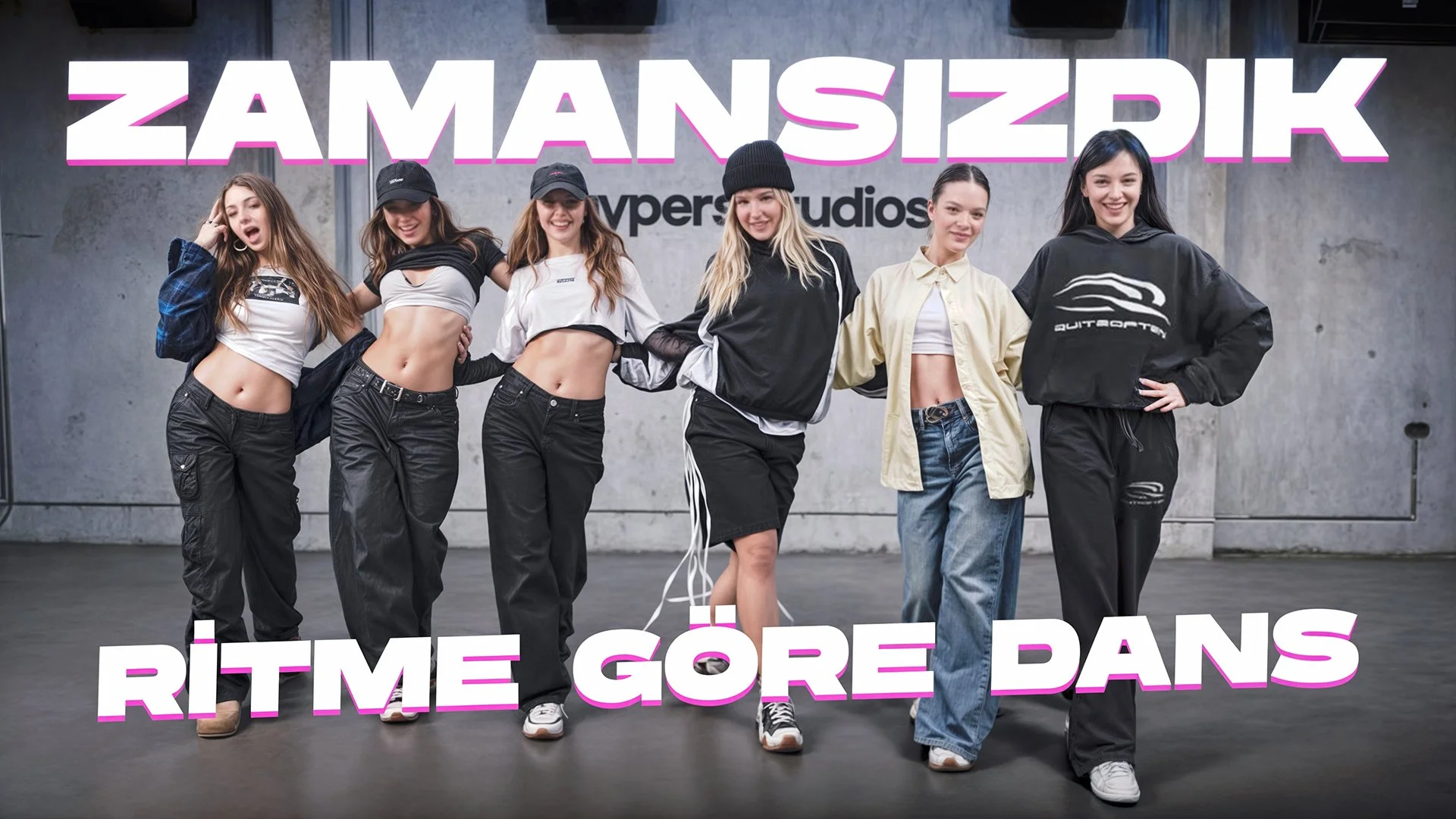 Six young women pose together in a dance studio, wearing casual sporty clothing, with a large sign reading 'ZAMANSIZIK RITME GÖRE DANS' in the background.