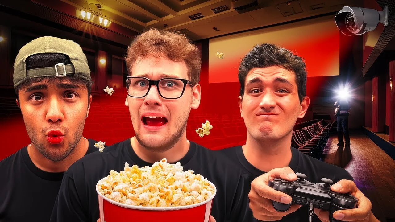 Three young men in a movie theater with empty seats, one holding popcorn, another with a game controller, and a third making a surprised face, with security cameras visible in the background.