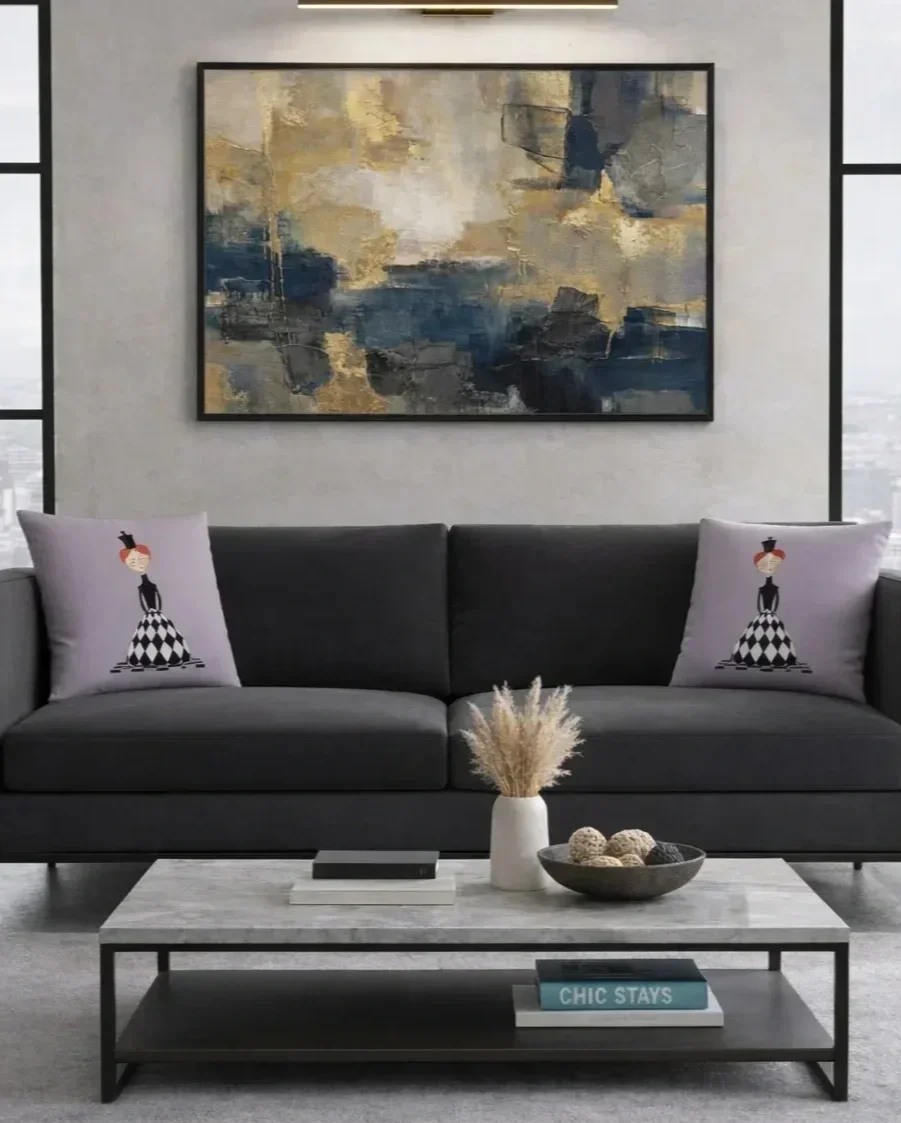 Modern living room with a dark gray sofa, two decorative pillows with illustrated women in black and white checkered dresses, a marble-top coffee table with a plant in a white vase, a bowl of decorative balls, and a large abstract painting with shades of blue, beige, and gray on the wall.