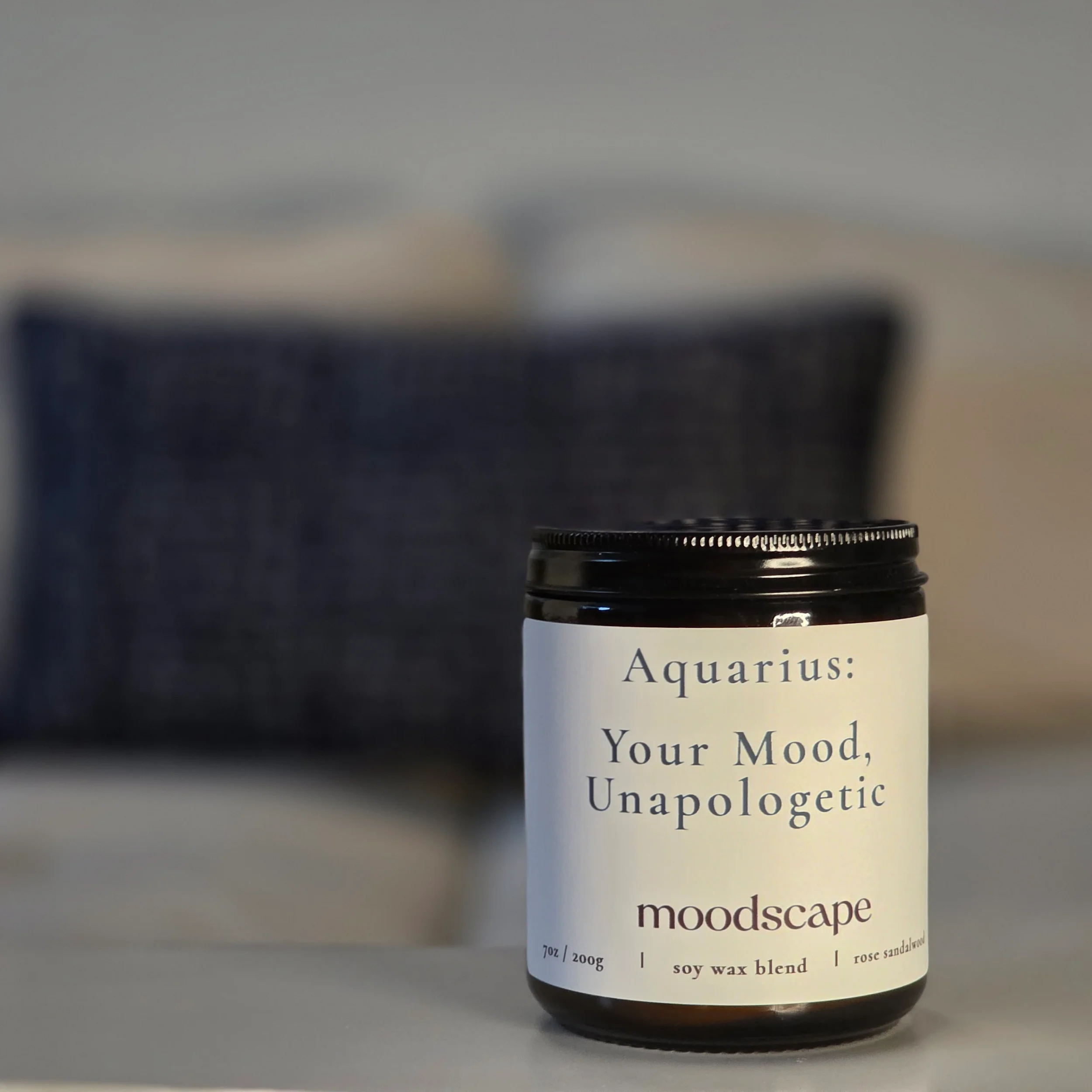 A jar with a black lid labeled 'Aquarius: Your Mood, Unapologetic.' The label includes additional text indicating it is a soy wax blend with rose sandalwood, weighing 7 ounces or 200 grams. The background is blurred.