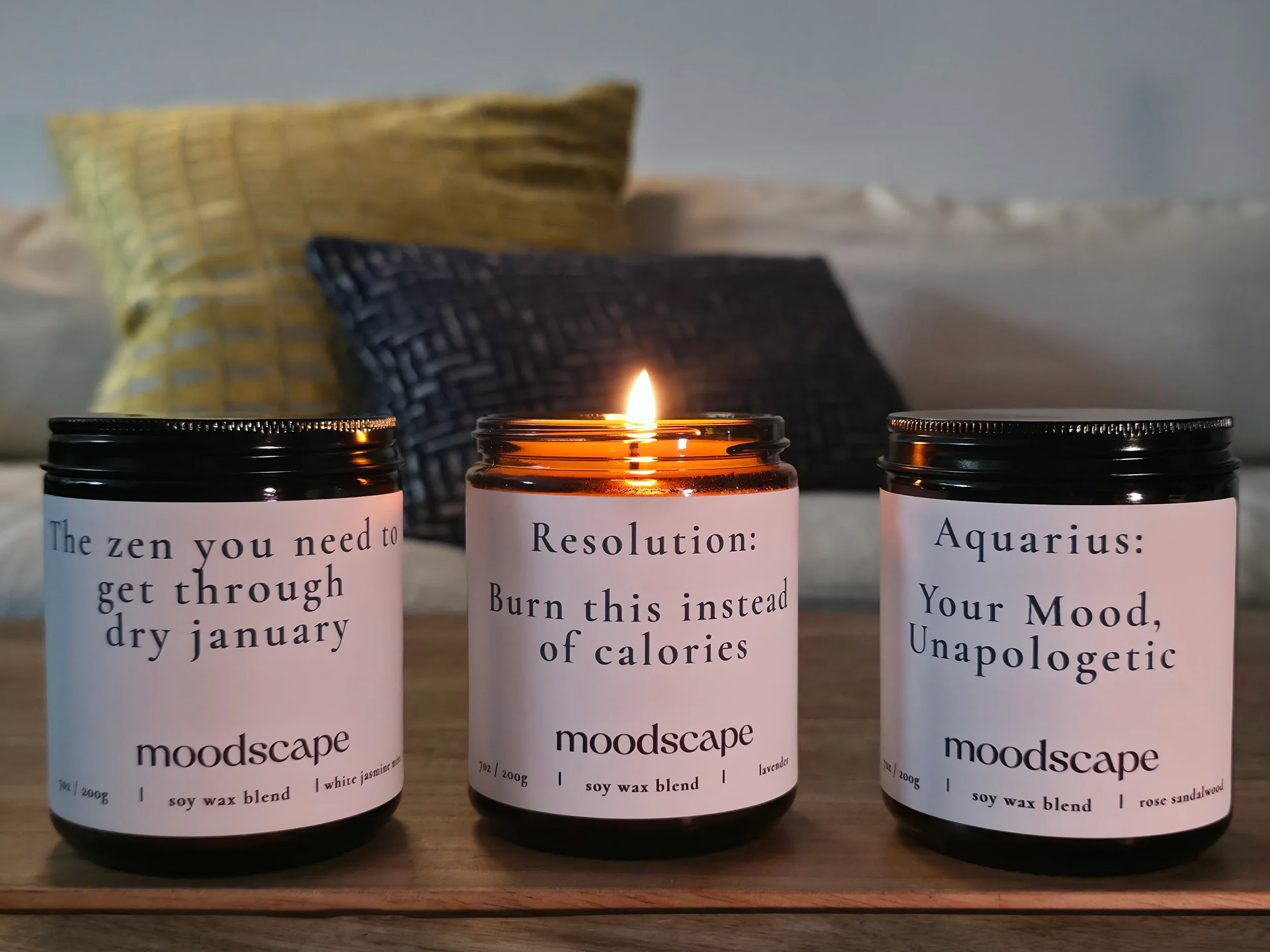 Three candles on a wooden tray, with pillows in the background. The middle candle is lit, and the jars are labeled with different wellness messages and scents.