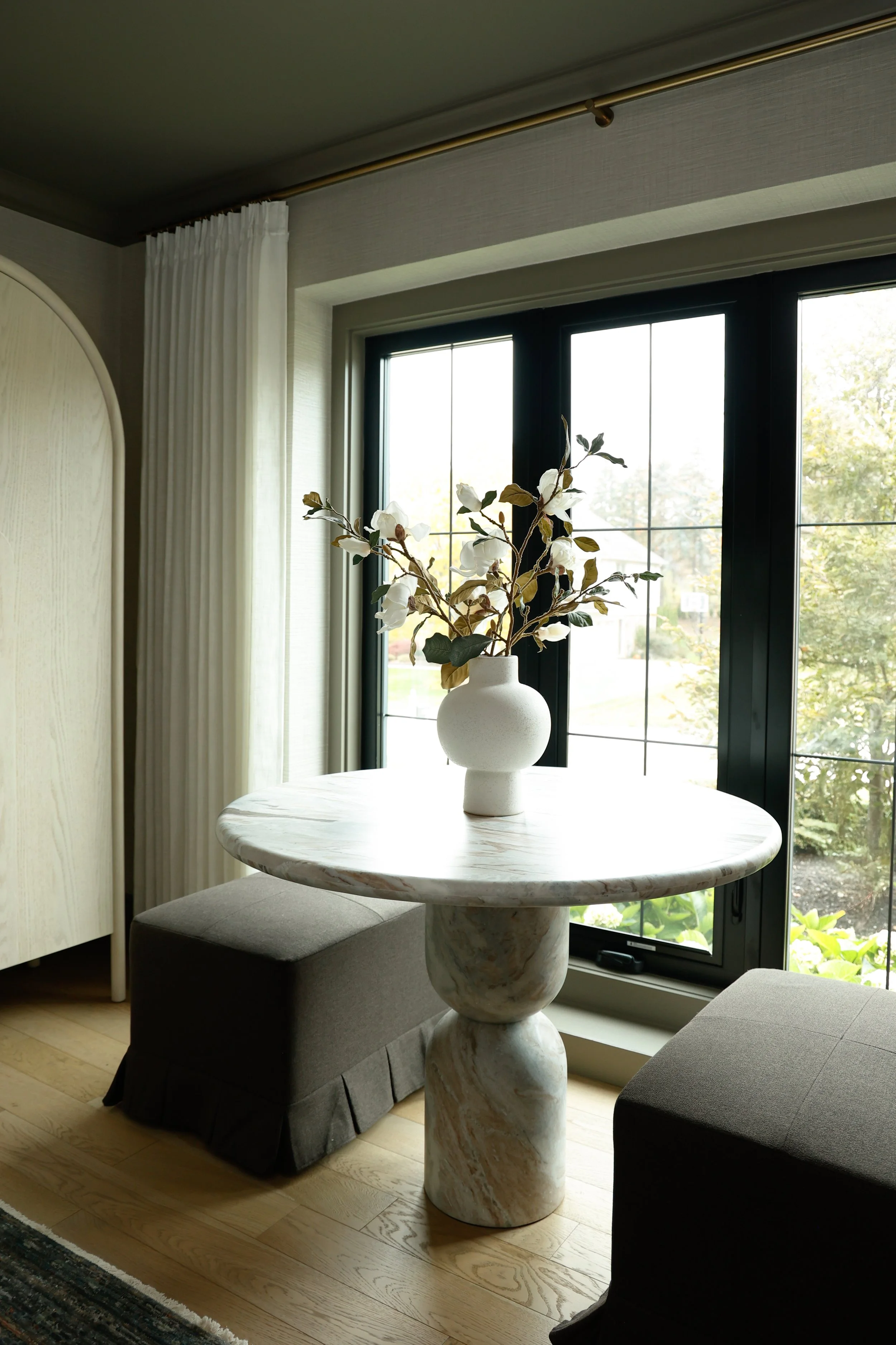 A round marble table with a white vase containing white flowers and green leaves, placed beside a window with white curtains in a cozy room.