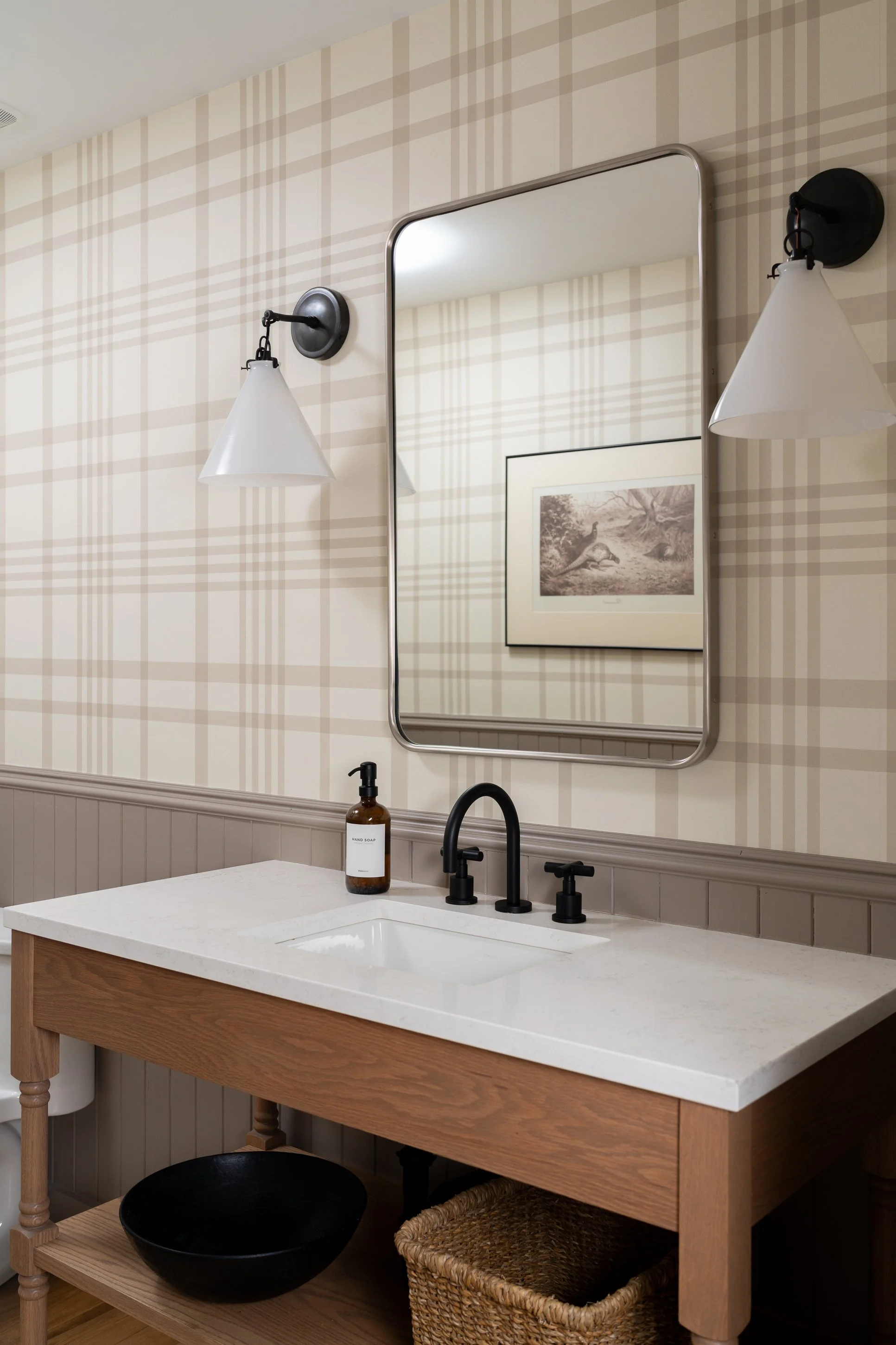 Bathroom vanity with a white countertop, black faucet, mirror, framed artwork, wall-mounted sconces, and a bottle of hand soap.