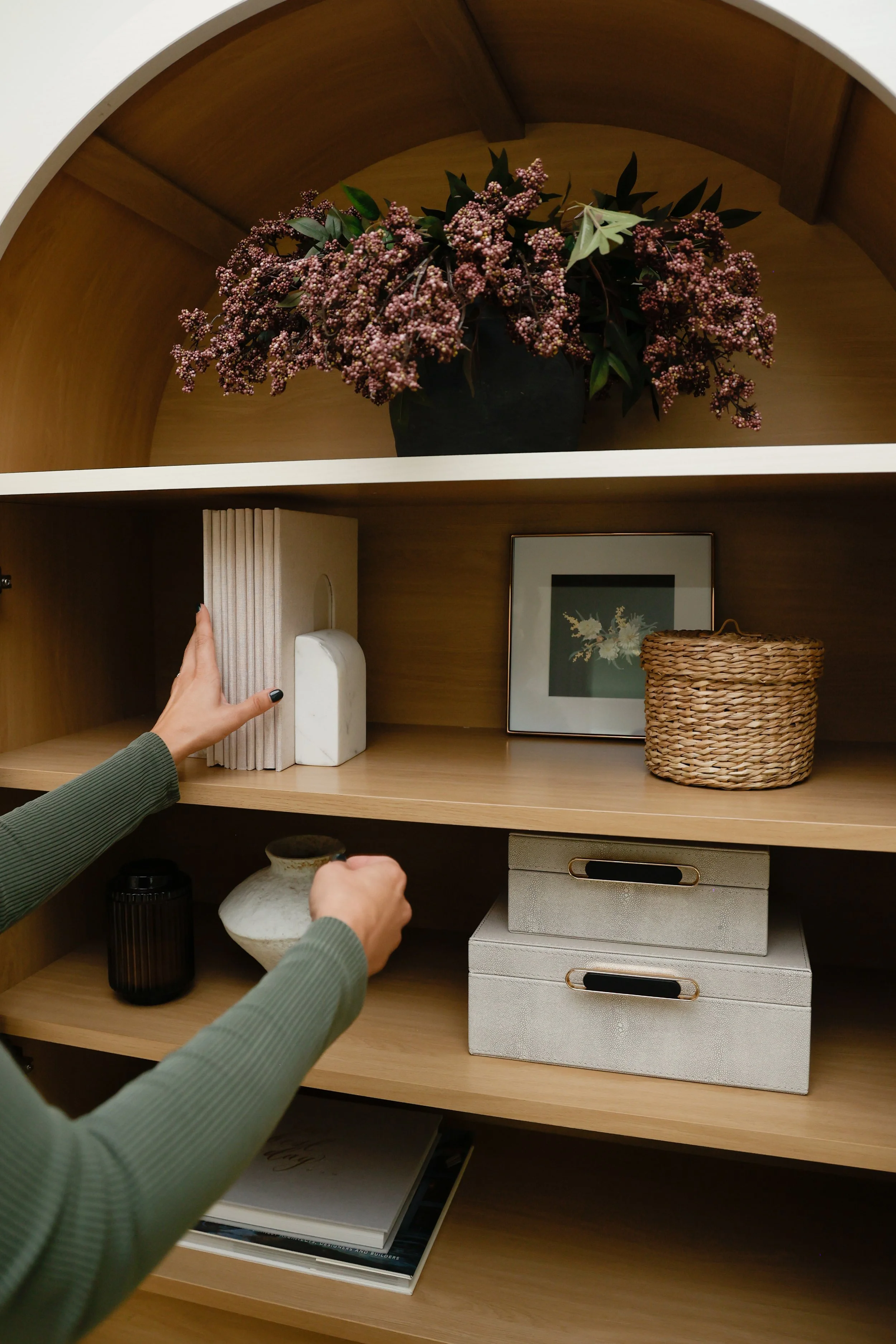A wooden bookshelf with two shelves containing various decorative items. The top shelf has a black vase with pink flowers and green leaves. The middle shelf has several books, a white marble block, a framed picture of flowers, a woven basket, and two white storage boxes. A hand with black nail polish is placing a book on the middle shelf, and another hand is reaching for a different item.