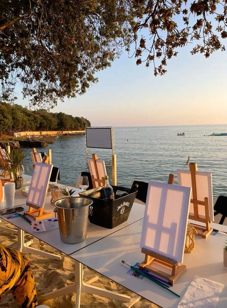 Outdoor beach scene with rectangular tables set up, featuring small canvas easels, wine bottles, glasses, and art supplies, with a view of the ocean, boats, and beach in the background at sunset.