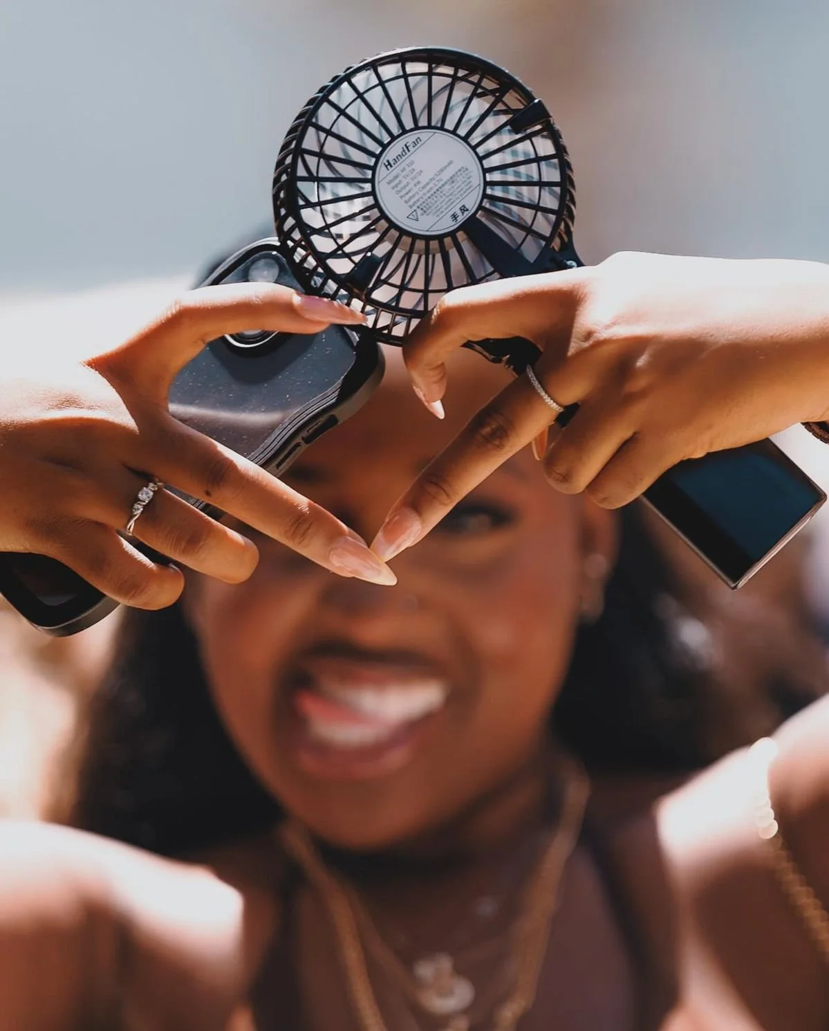 A smiling woman makes a heart shape with her hands around a small portable fan, with her face blurred in the background.