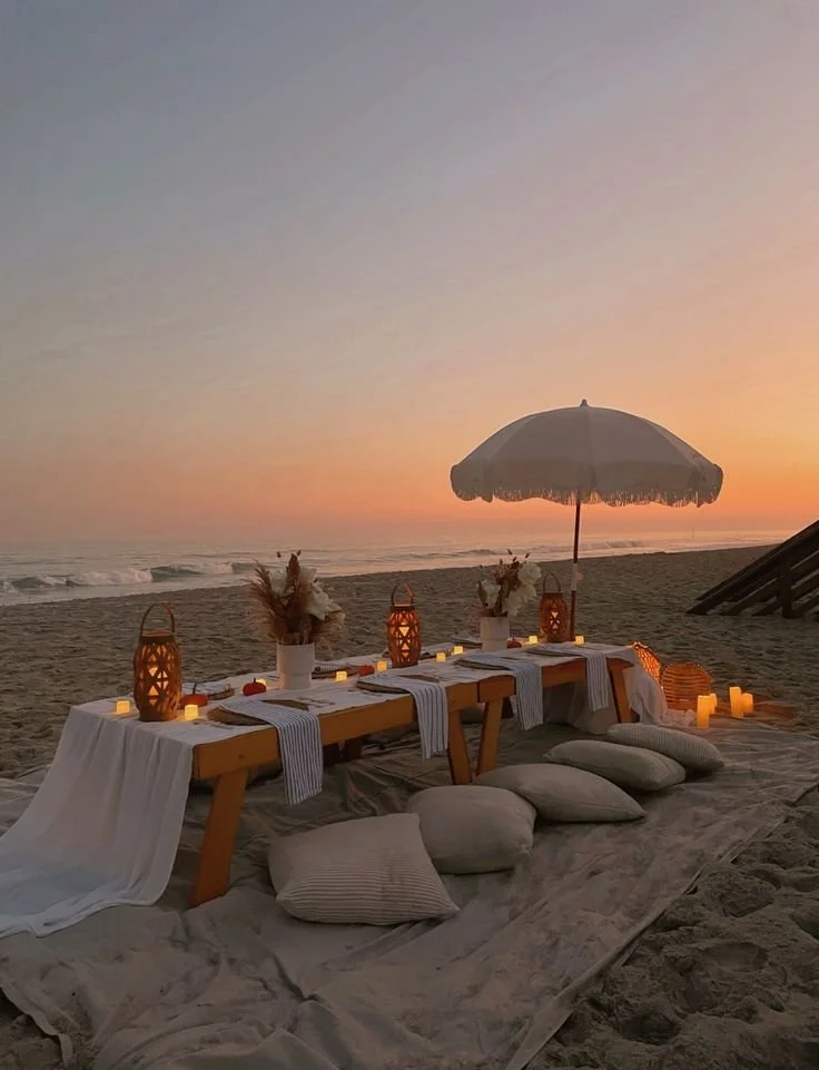 Beachside dining setup with a long table, pillows on the sand, and a large umbrella during sunset, illuminated by candles and lanterns.