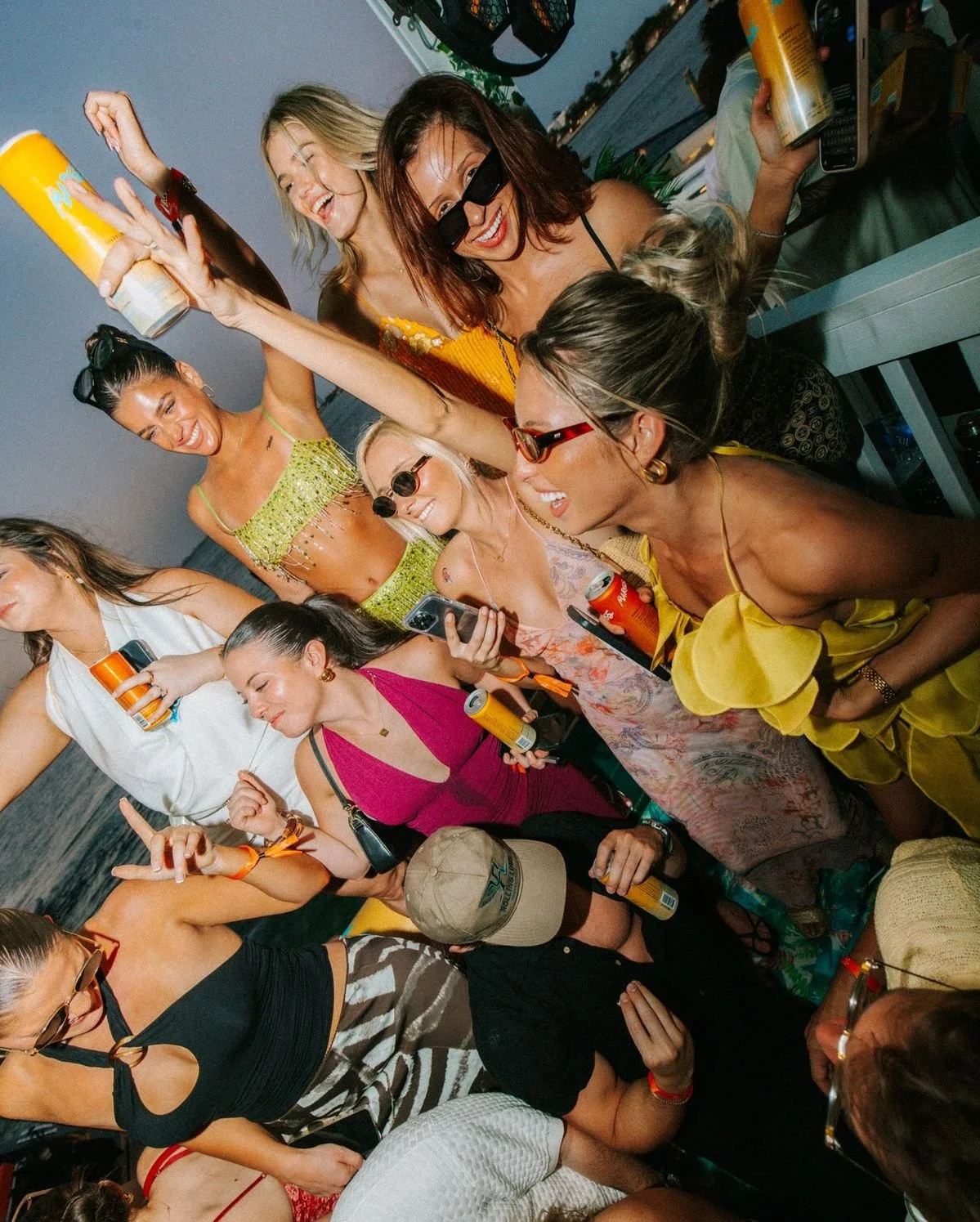 Group of women celebrating, holding drinks, smiling, and taking photos during a party or gathering by the water at sunset.