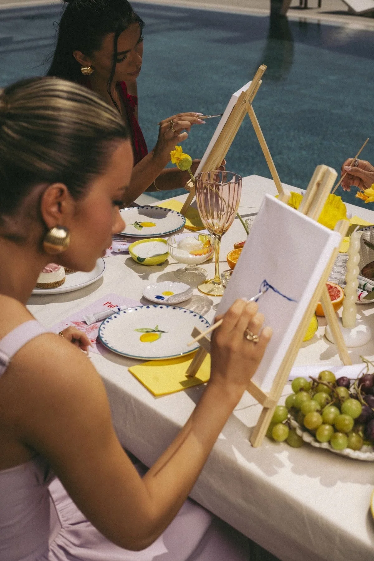 Two women painting on canvases set on small wooden easels at an outdoor table near a pool, with snacks and drinks around them, during evening.