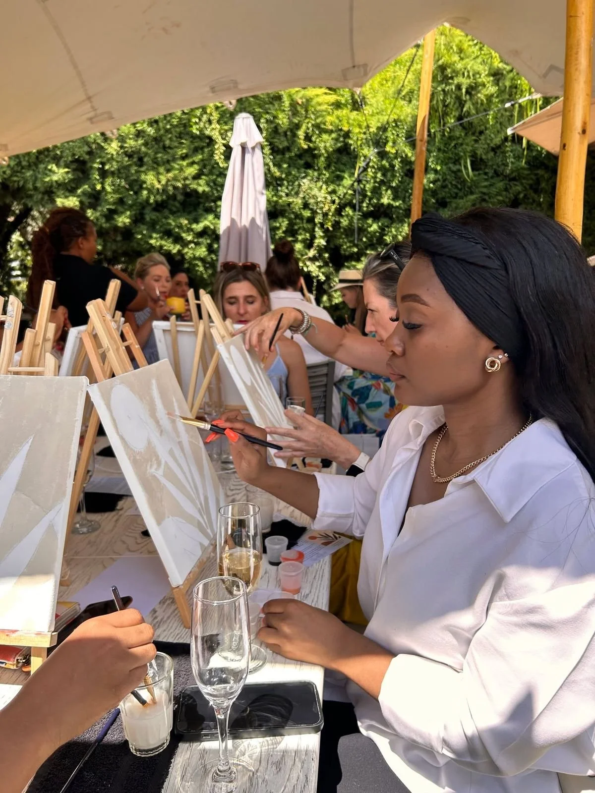 Group of women participating in a painting class outdoors, sitting at a table with canvases and paints under a canopy, with greenery in the background.