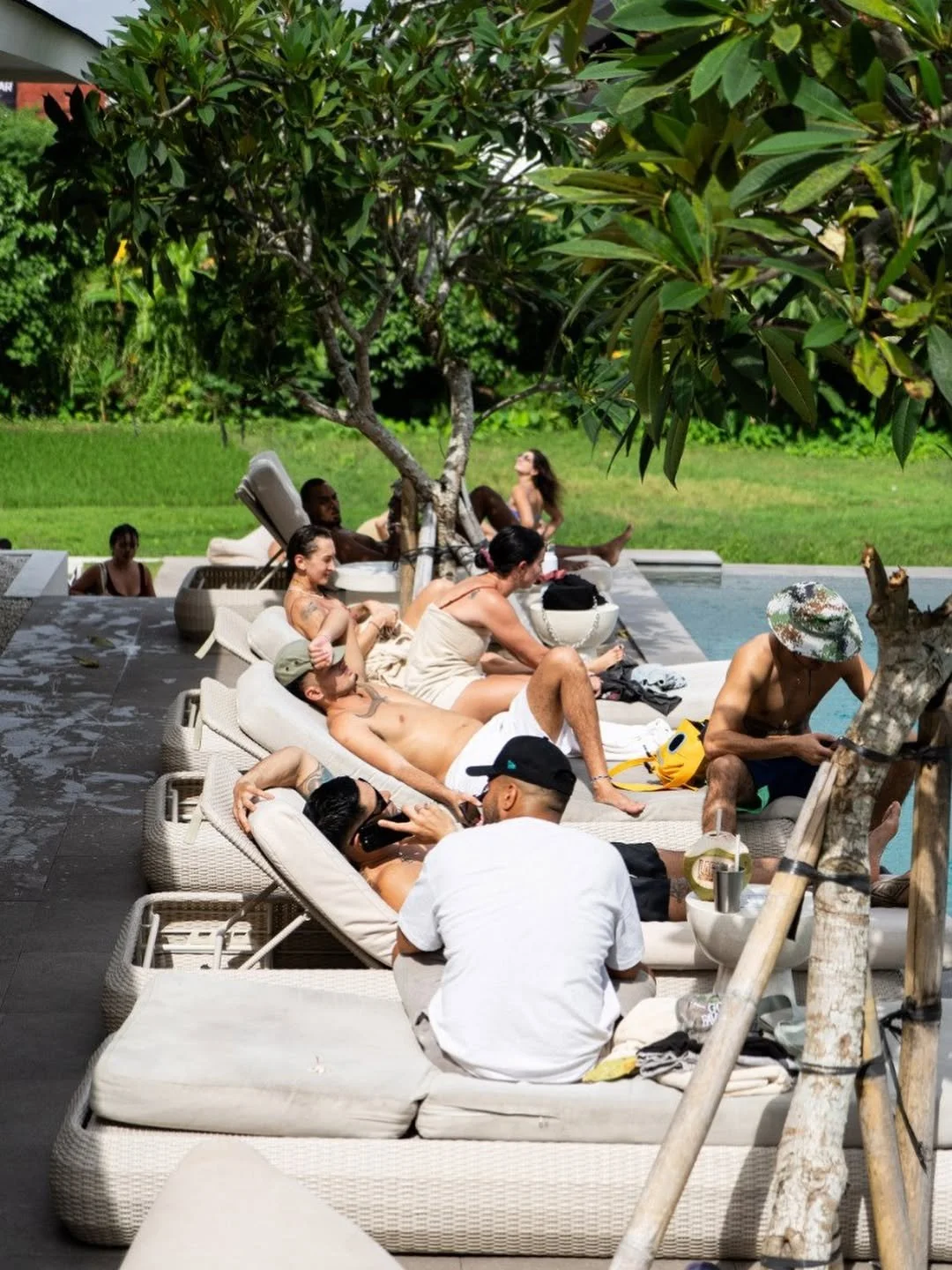People relaxing on outdoor lounge chairs and enjoying a sunny day by a pool, with trees and greenery in the background.