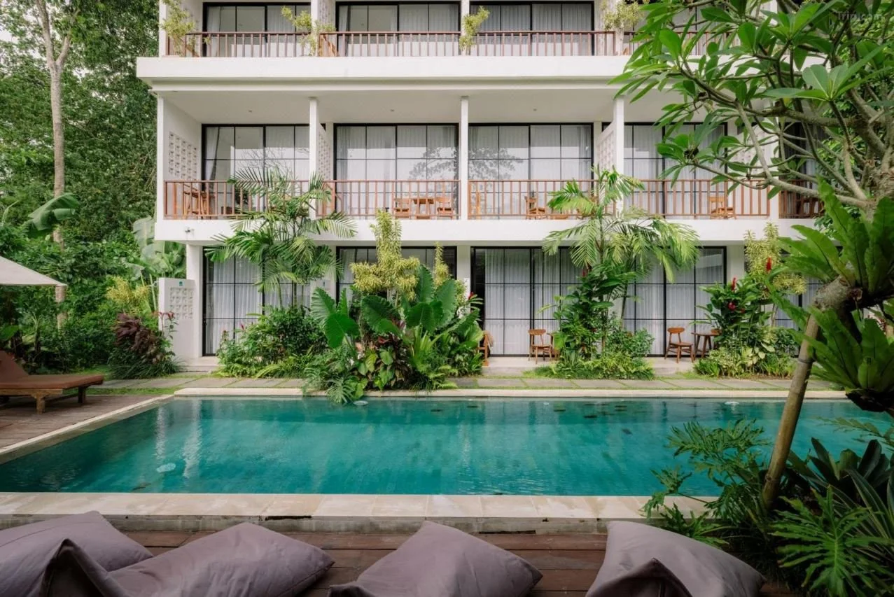 A modern hotel or apartment building with balconies, surrounded by lush green plants, with a swimming pool in the foreground, and lounge chairs and cushions nearby.