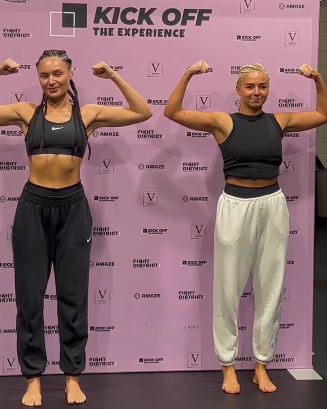 Two women standing on a black floor, flexing their biceps in front of a pink backdrop with logos and the text 'Kick Off The Experience'. The woman on the left is wearing a black sports bra and black sweatpants, with her hair in braids. The woman on the right is wearing a black sleeveless top and white sweatpants, with her hair in a braid. Both women are barefoot.