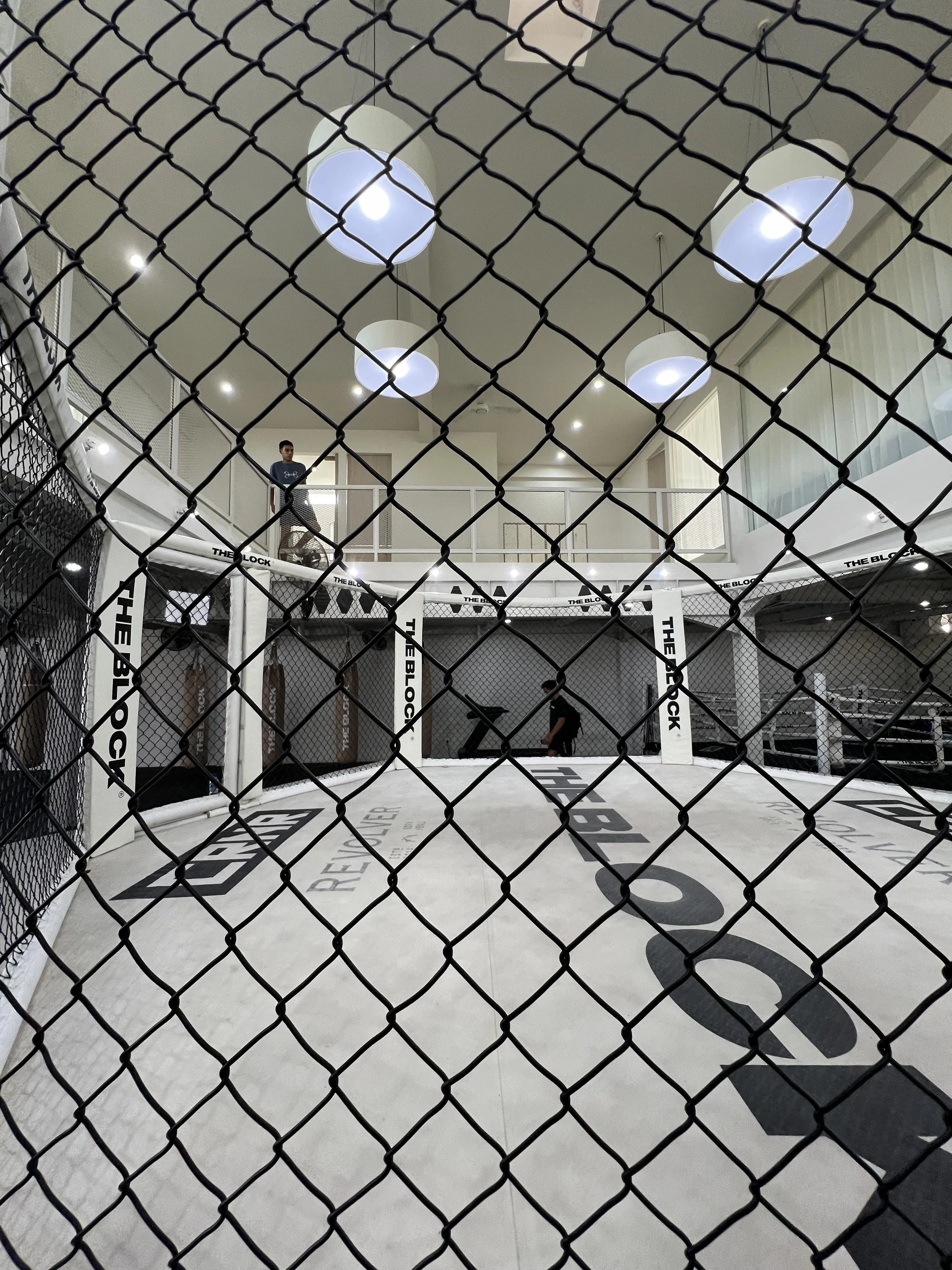 An indoor MMA or boxing training cage with black chain-link fencing, a white padded floor with black logos, and a person practicing inside. The cage is surrounded by a balcony with a person observing from above.