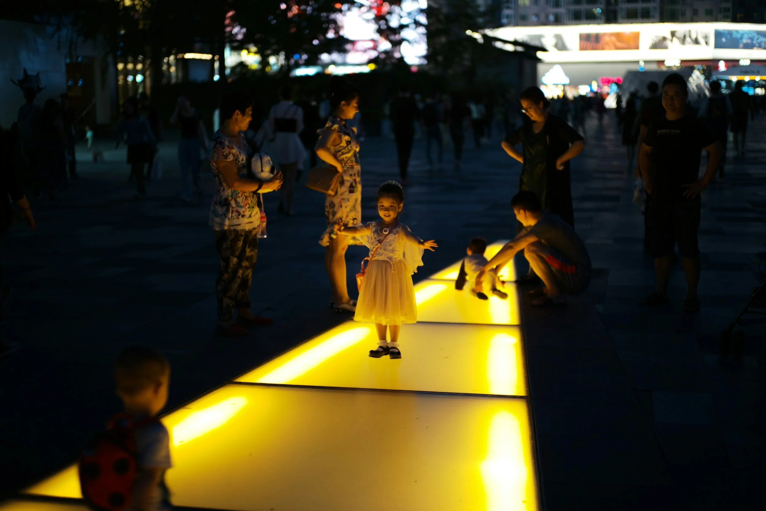 Children playing on illuminated yellow walkway at night with adults and other people in the background.