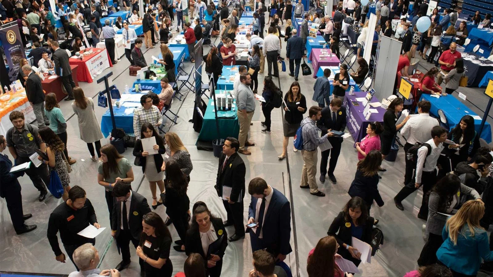 A busy job fair or career expo with multiple booths and many people walking, talking, and holding brochures or documents.