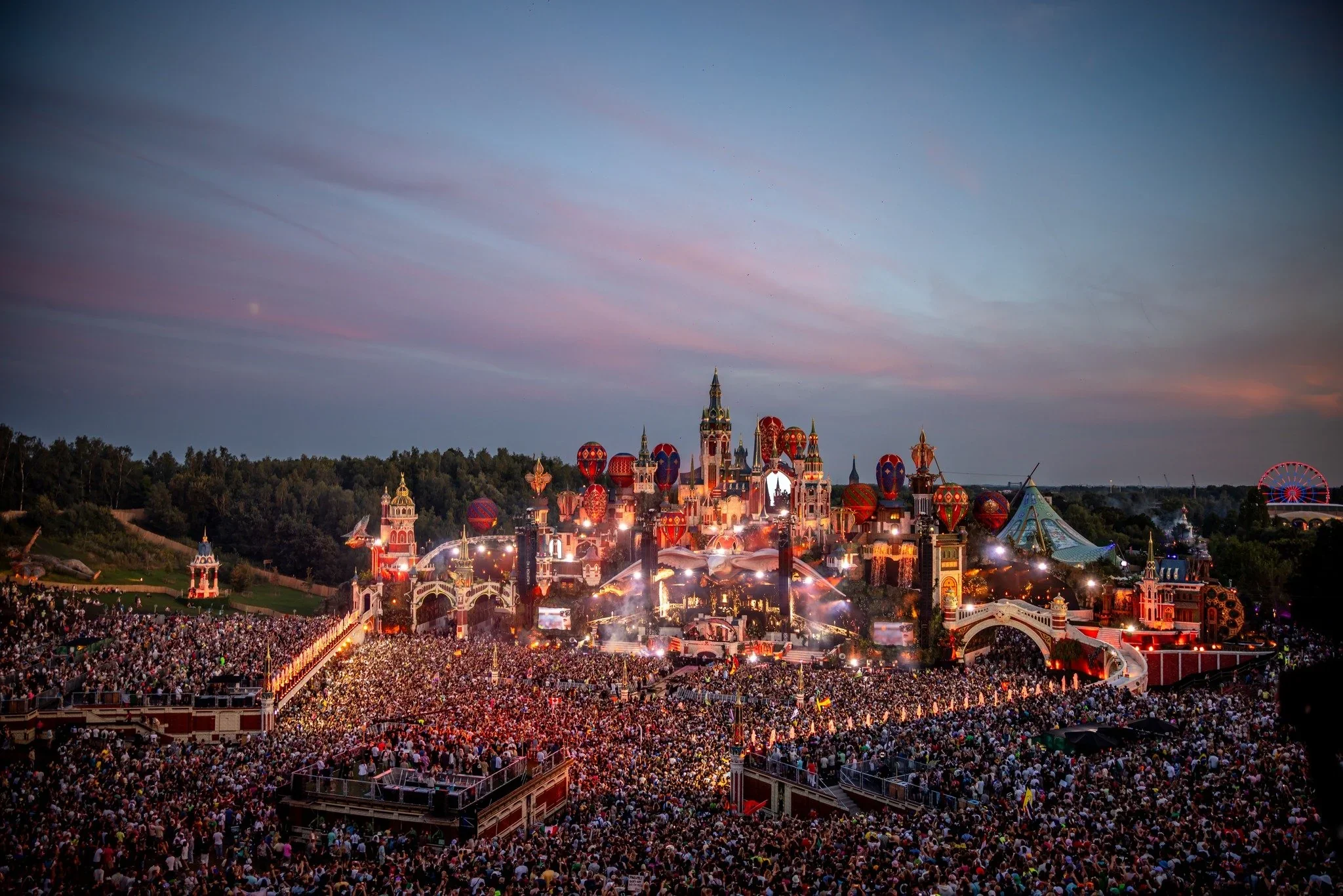 Nighttime view of a large crowd at a theme park with a castle illuminated with colorful lights and fireworks in the sky.
