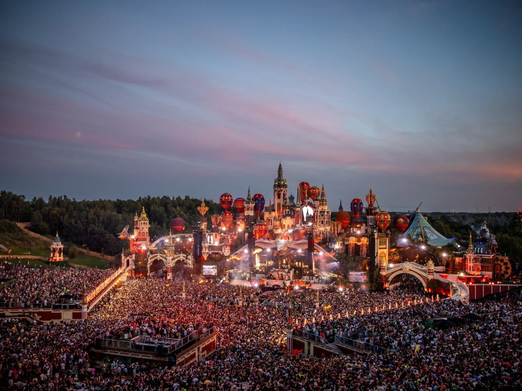 A large crowd gathered at a Disney theme park during sunset, with colorful castle and hot air balloons overhead.