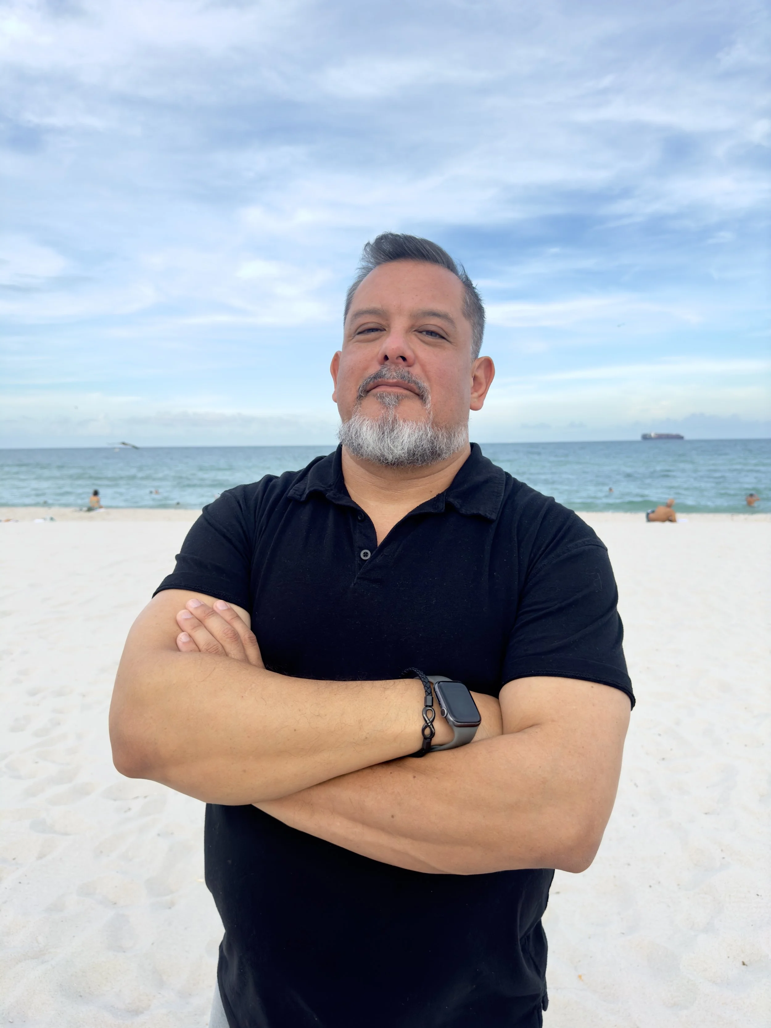 A man with crossed arms, wearing a black polo shirt and smartwatch, posing confidently on a beach with a blue sky, ocean, and other beachgoers in the background.