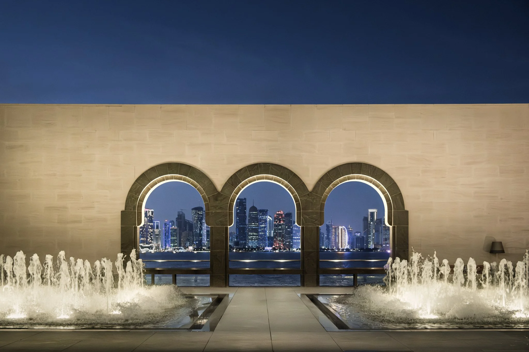 Architectural photography of the Museum of Islamic Art waterfront in Doha, Qatar. Three illuminated arches framing the West Bay skyline at night, with water fountains in the foreground.