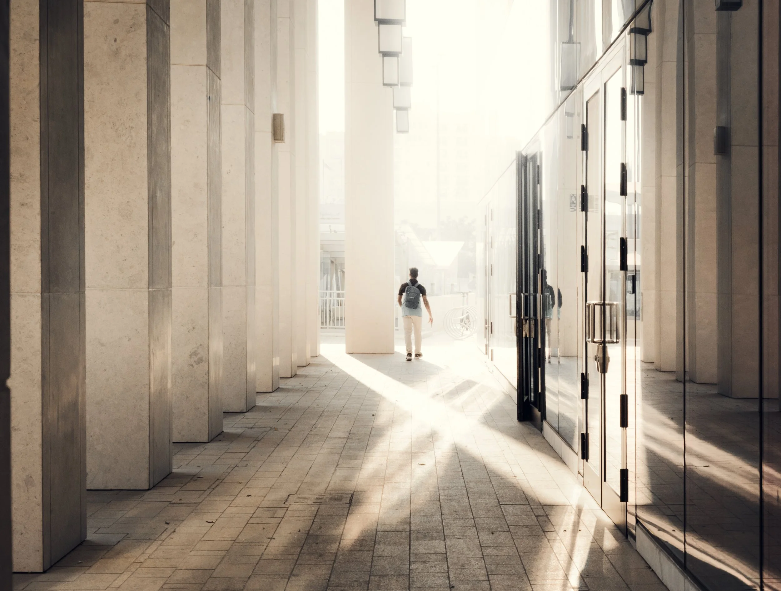 Urban passage in Msheireb Downtown Doha with strong directional light, vertical stone columns, reflective surfaces, and a pedestrian used as scale reference in architectural photography.