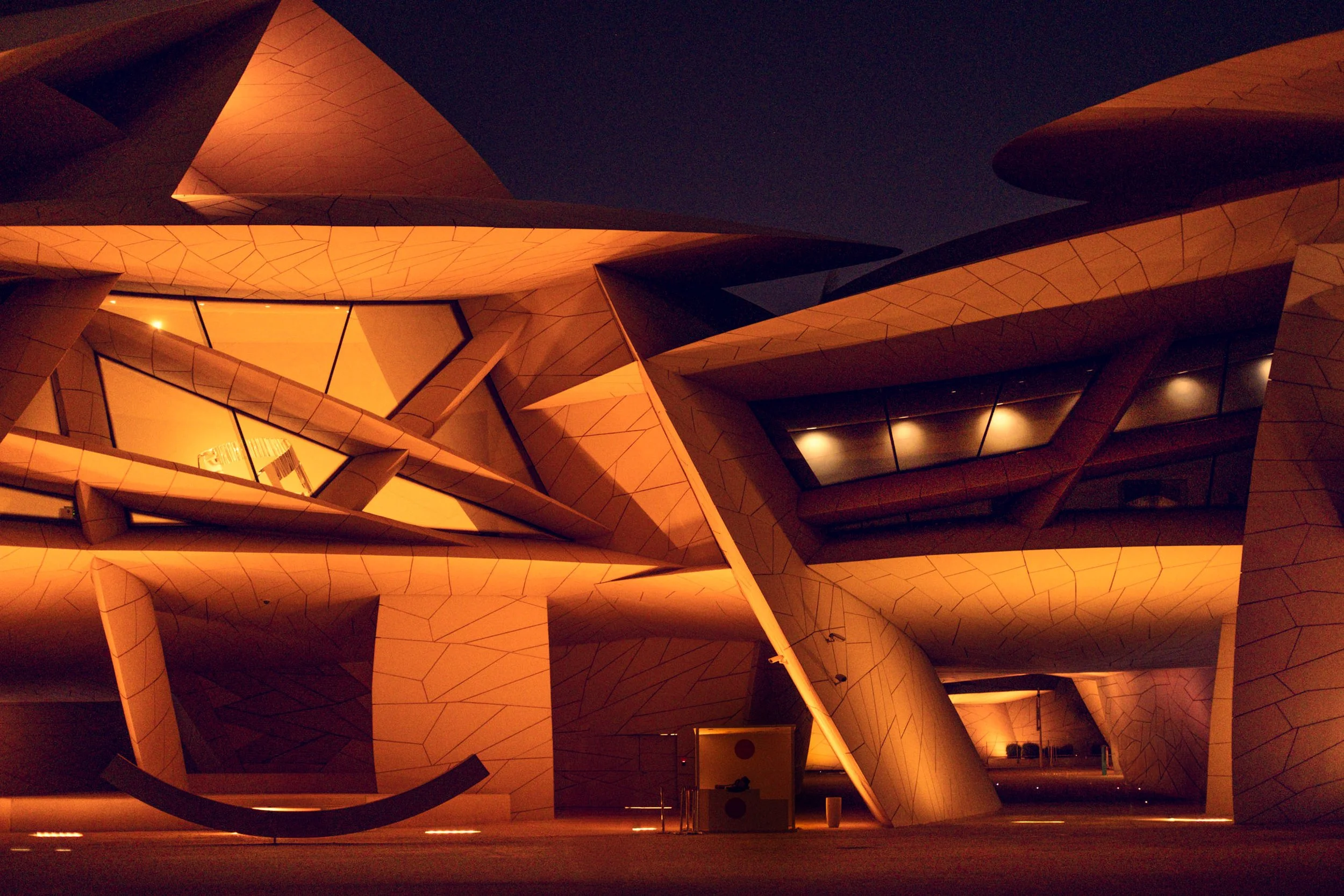 Night view of the National Museum of Qatar with warm light across the interlocking geometric plates, showing texture, sharp shadows and the building’s structure for architects and designers looking for references in Doha’s night architecture.