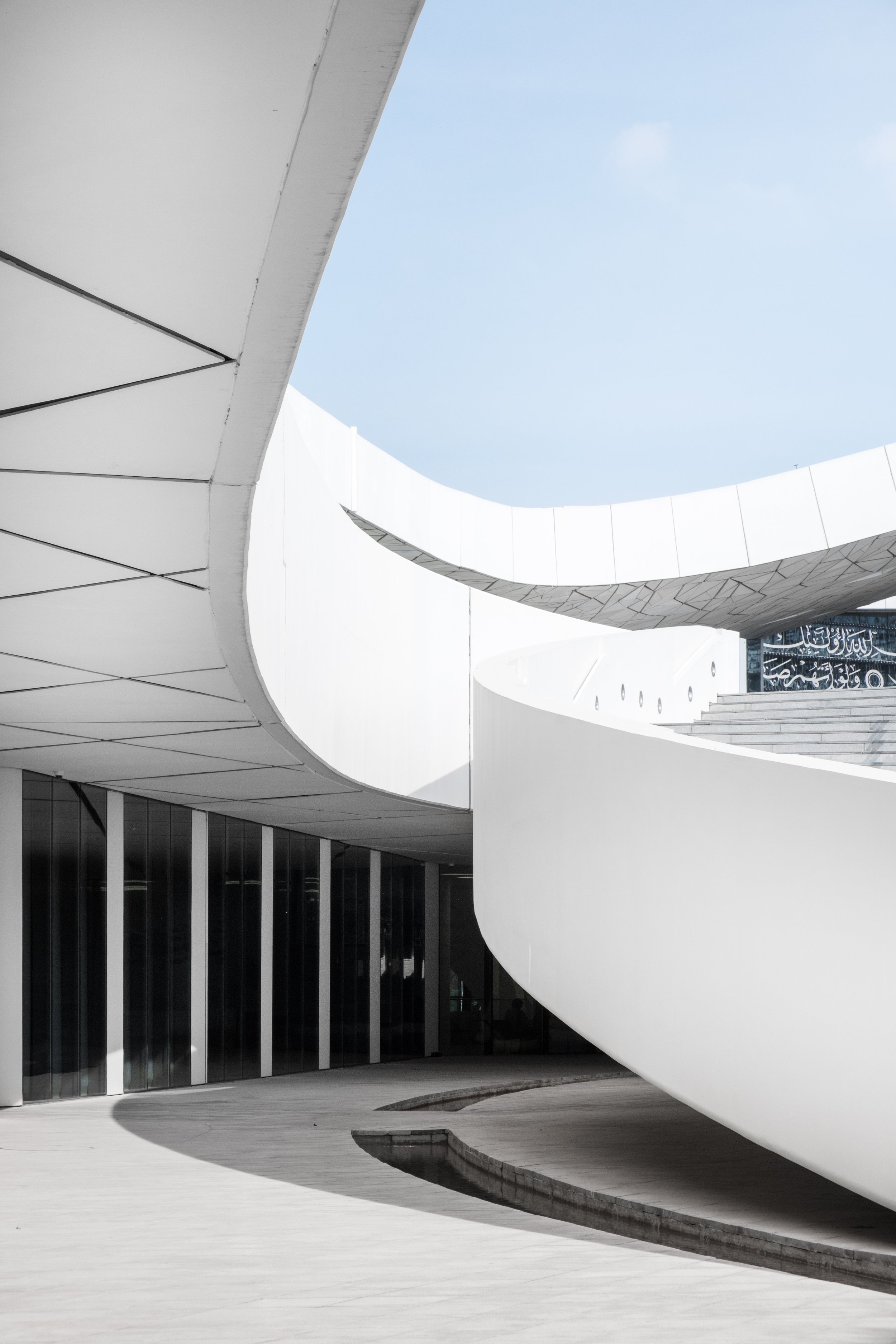 Low-angle architectural photography of the white stairs and sweeping curved facade of the Education City Mosque (Minaretein) in Doha, featuring intricate geometric tiling and Arabic calligraphy.