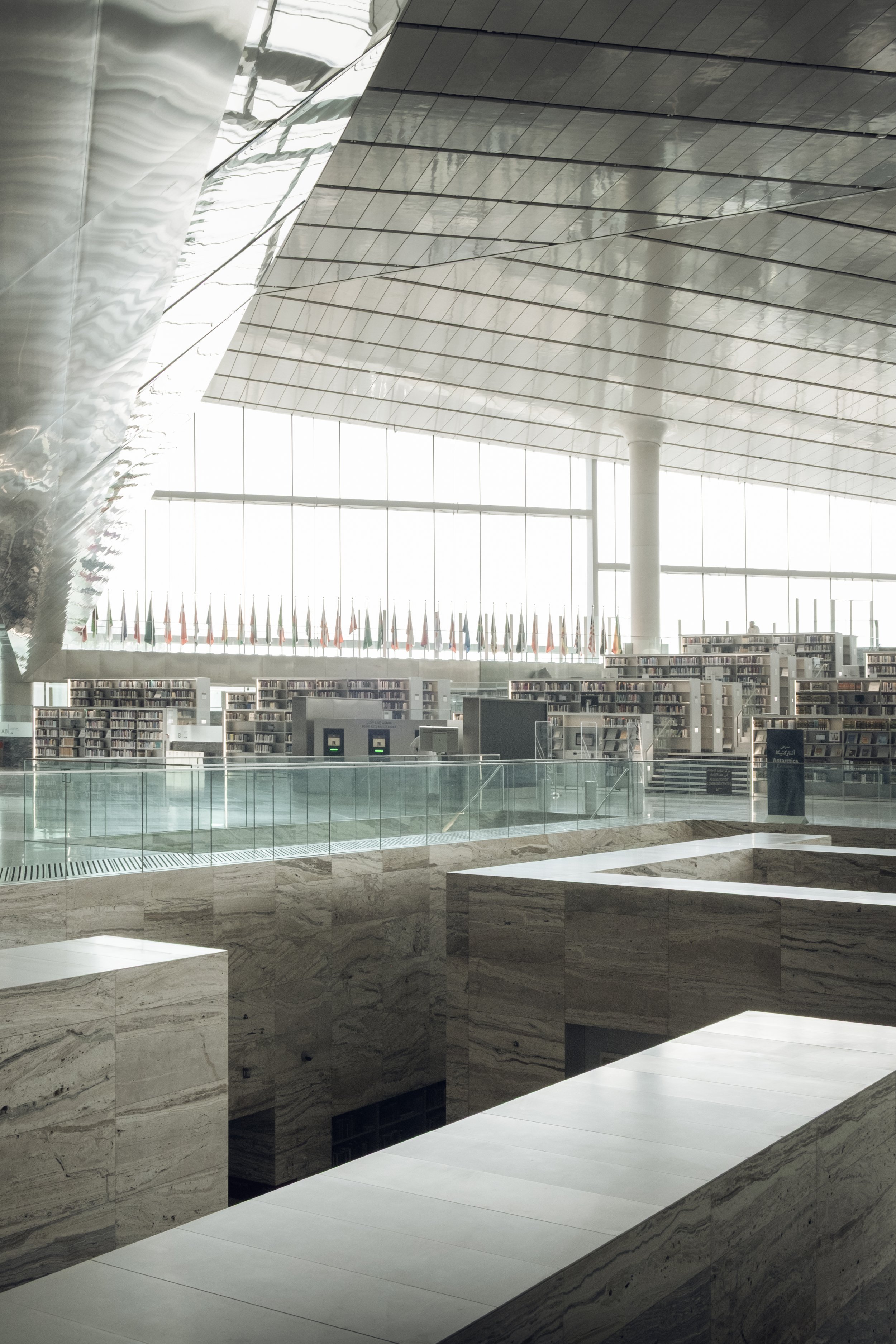 Interior architectural photograph of the Qatar National Library in Doha, showing layered bookshelves, marble platforms and glass bridges that create depth and scale, captured with balanced natural light for architects and interior designers.