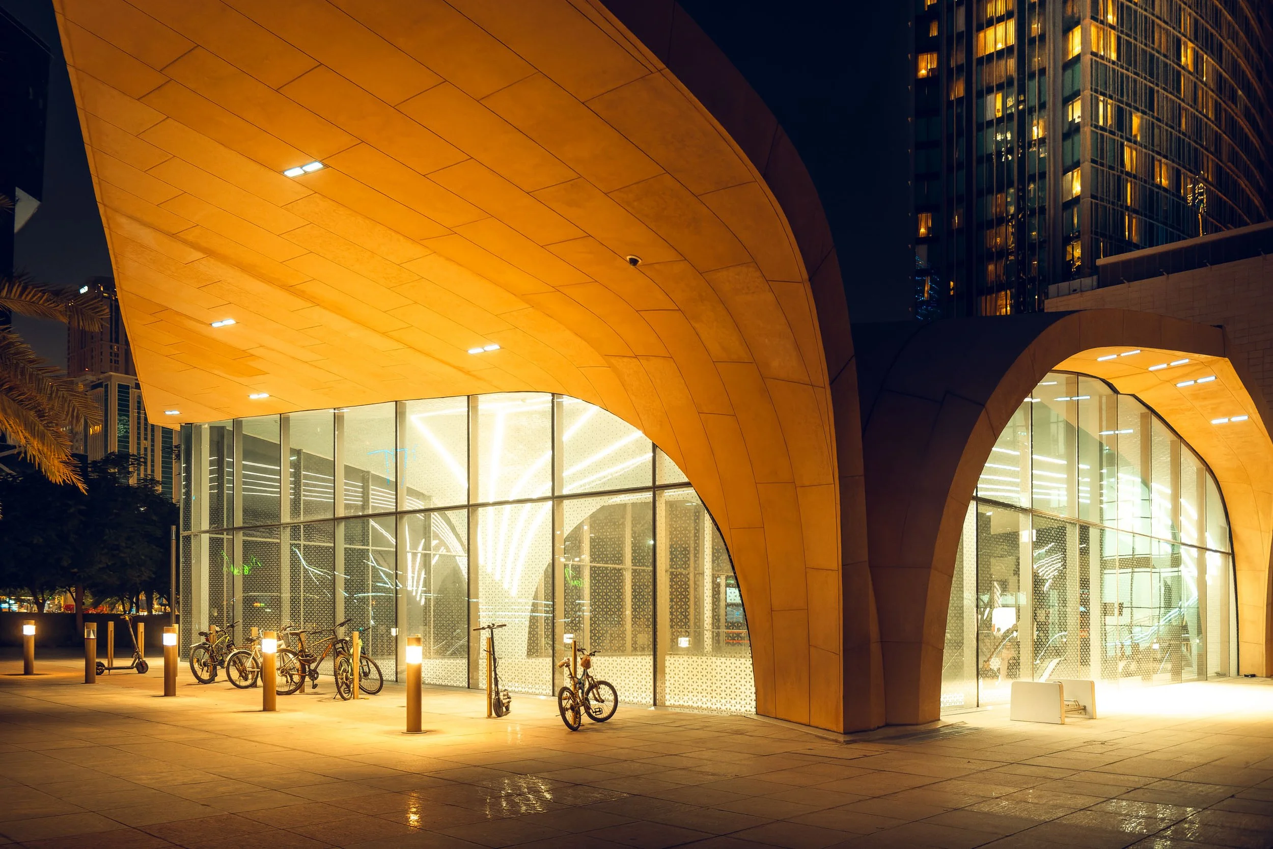 Curved entrance structure in West Bay at night with warm light across the concrete surface and bright interior lines visible through the glass facade, showing Doha’s blend of organic forms and modern lighting for architecture and design audiences.