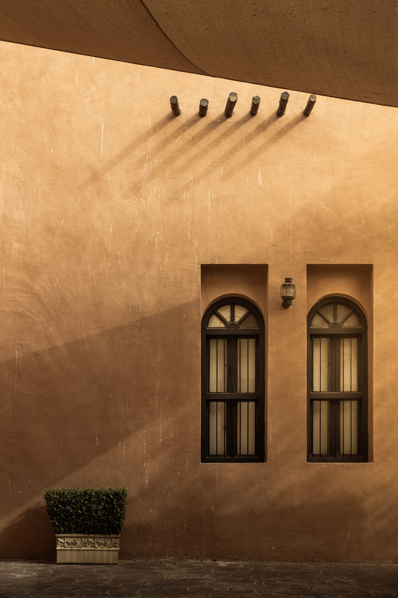 Architectural photography of traditional Qatari clay walls in Doha, showcasing dramatic diagonal shadows, two arched windows with dark frames, and wooden roof beams. Professional photography by Pedro Ferr