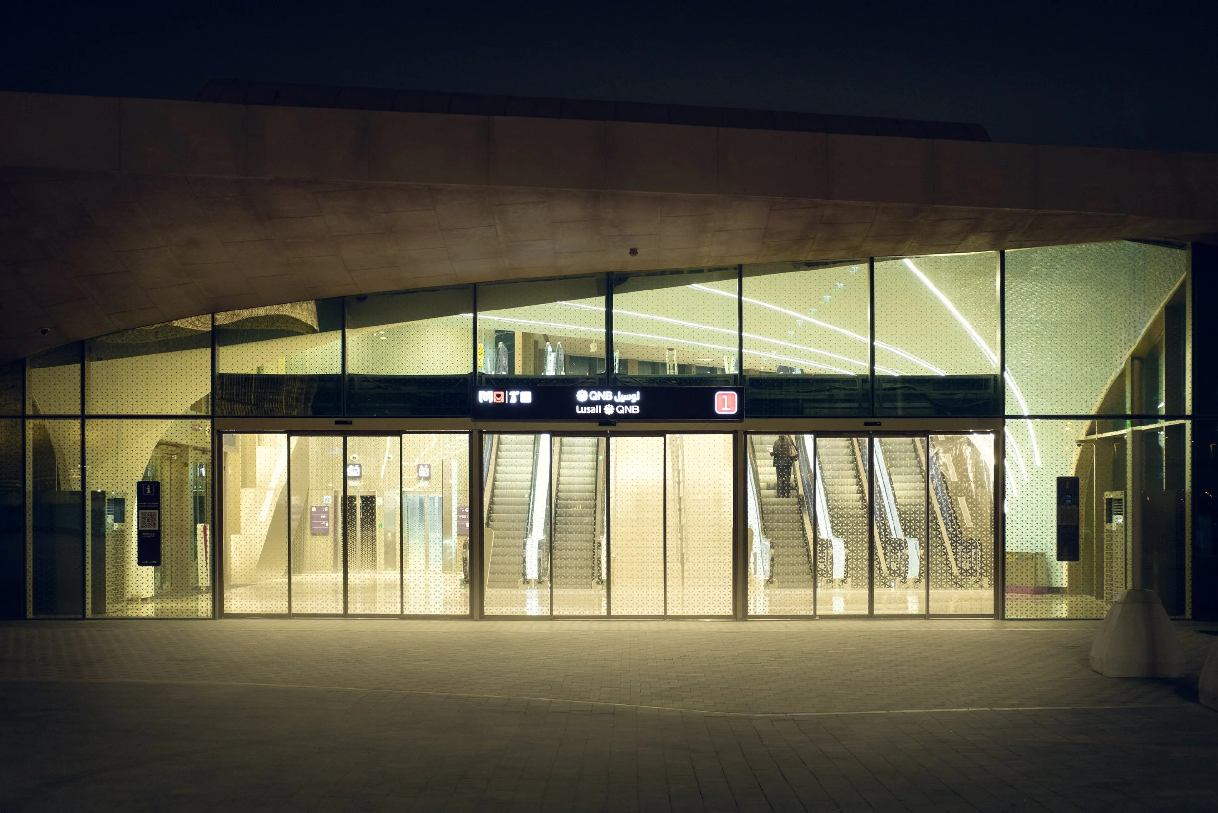 Glass facade of the Doha Metro Lusail QNB station at night with escalators illuminated inside, clean lines, soft warm light and curved forms that show how Doha uses controlled lighting in public architecture for design and architecture audiences.