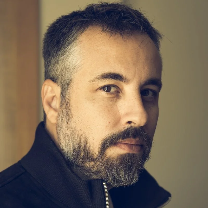 Close-up of a man with short dark hair, a beard, and mustache, wearing a black zip-up jacket, looking at the camera with a serious expression.