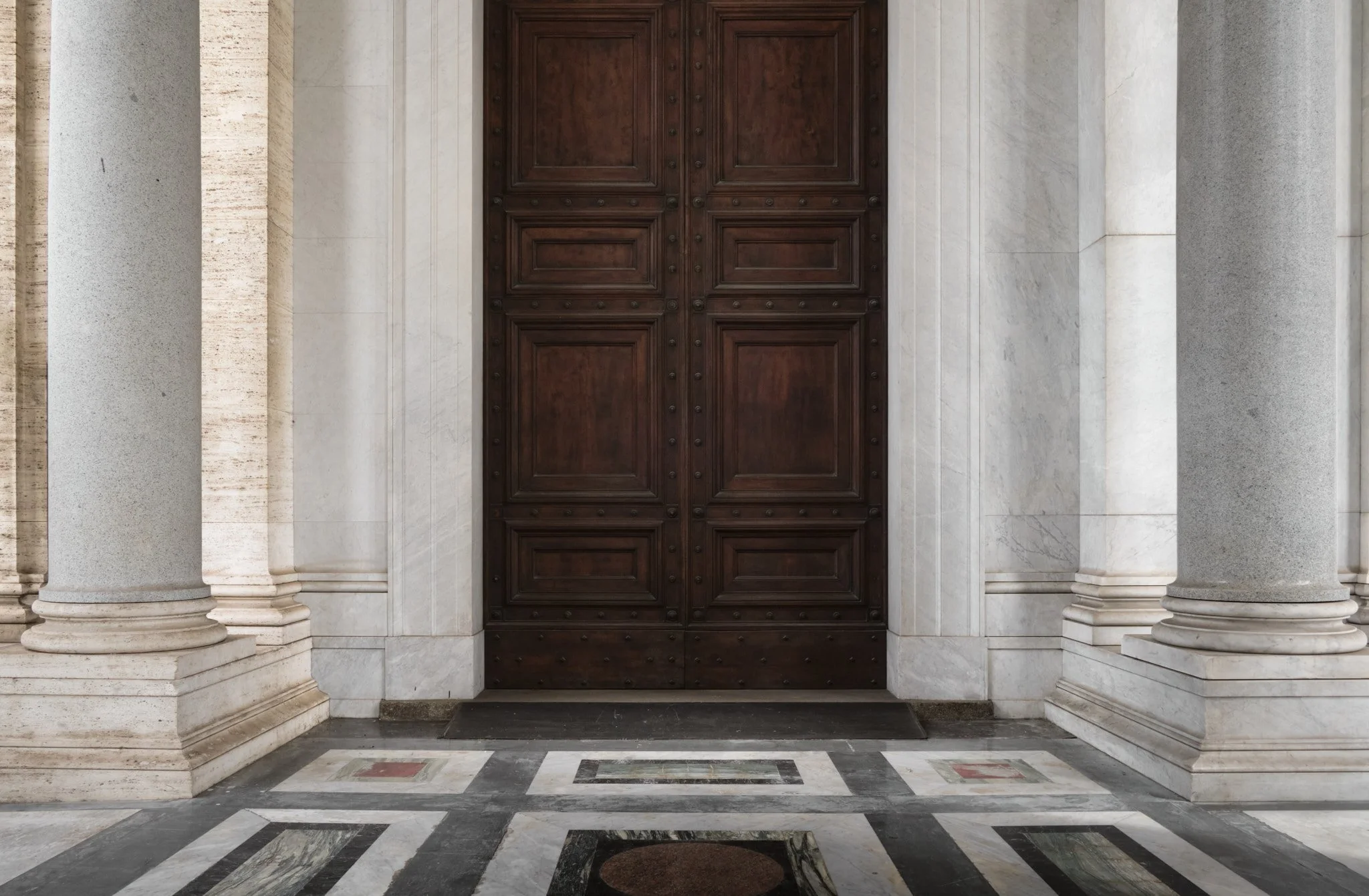 Centered composition of the bronze entrance doors of the Basilica di San Paolo Fuori le Mura, flanked by marble columns, marble threshold visible in available dark light.