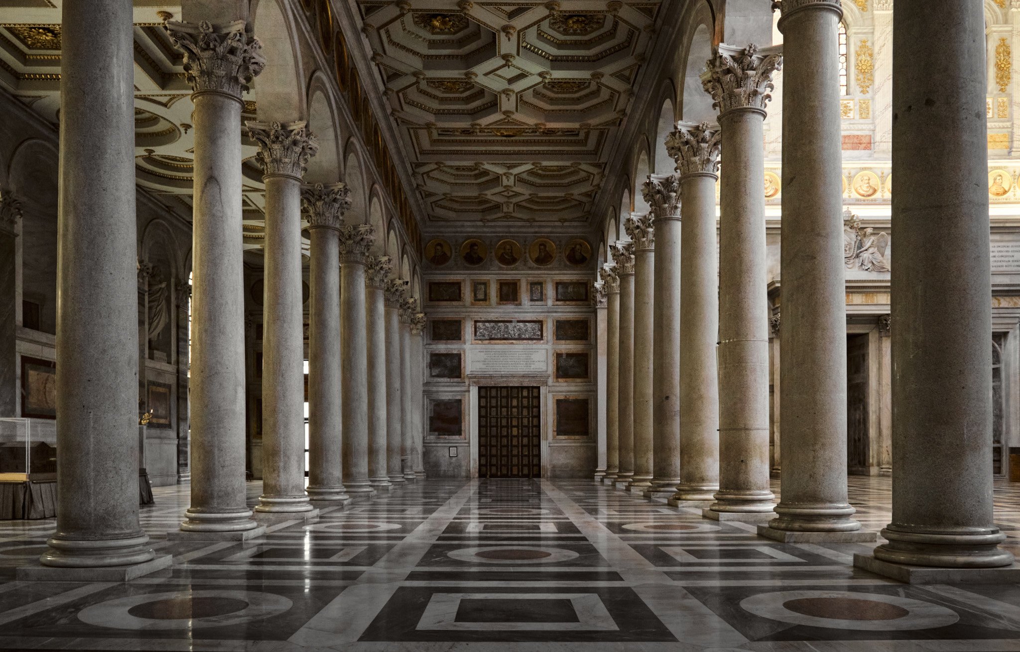 nterior colonnade of a side aisle at Basilica di San Paolo Fuori le Mura, dark marble floor with columns leading toward a bright lit exit or narthex beyond.
