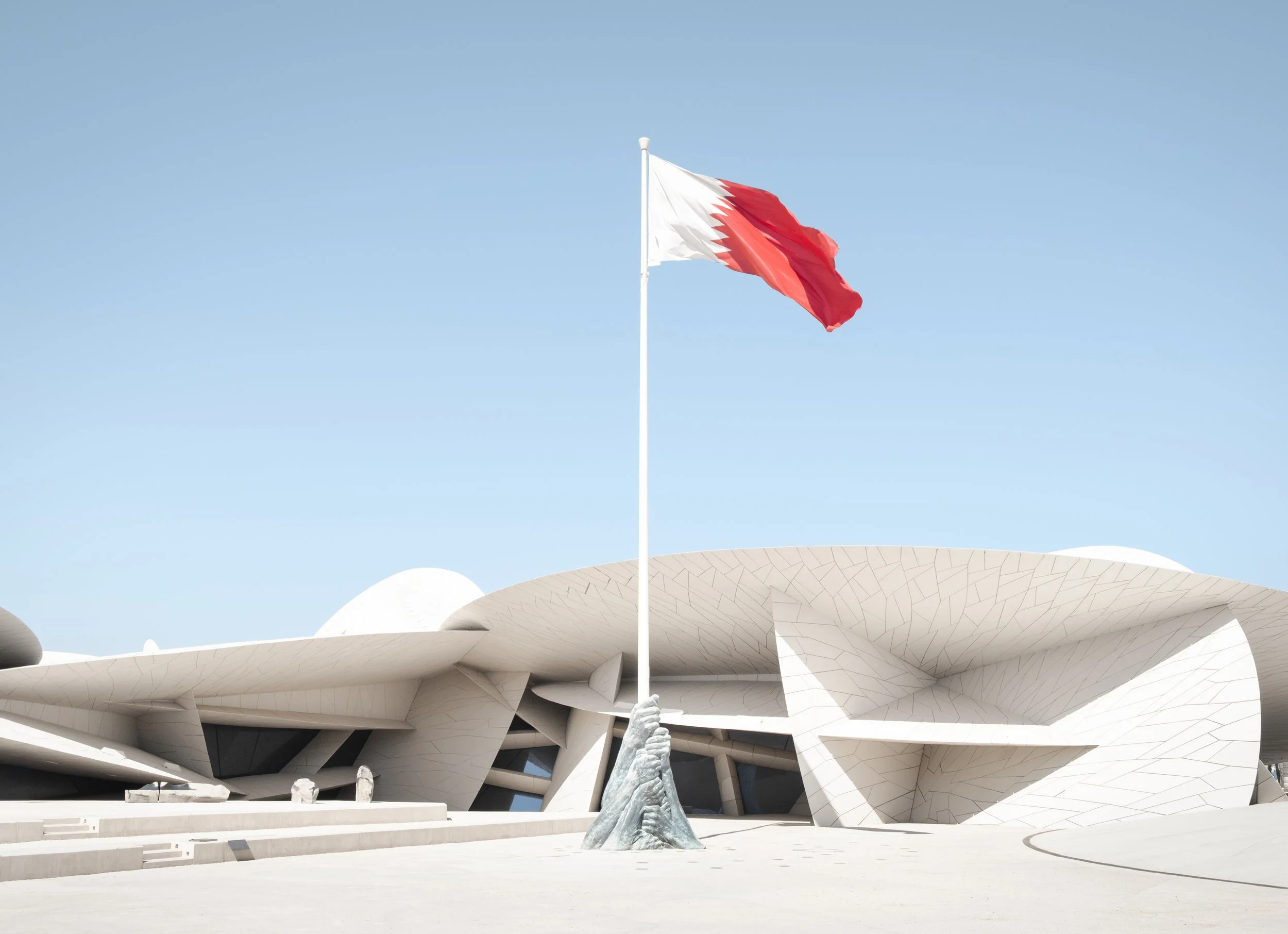 Qatari flag waving above the National Museum of Qatar in Doha, designed by architect Jean Nouvel and inspired by desert rose formations, photographed under clear desert light.