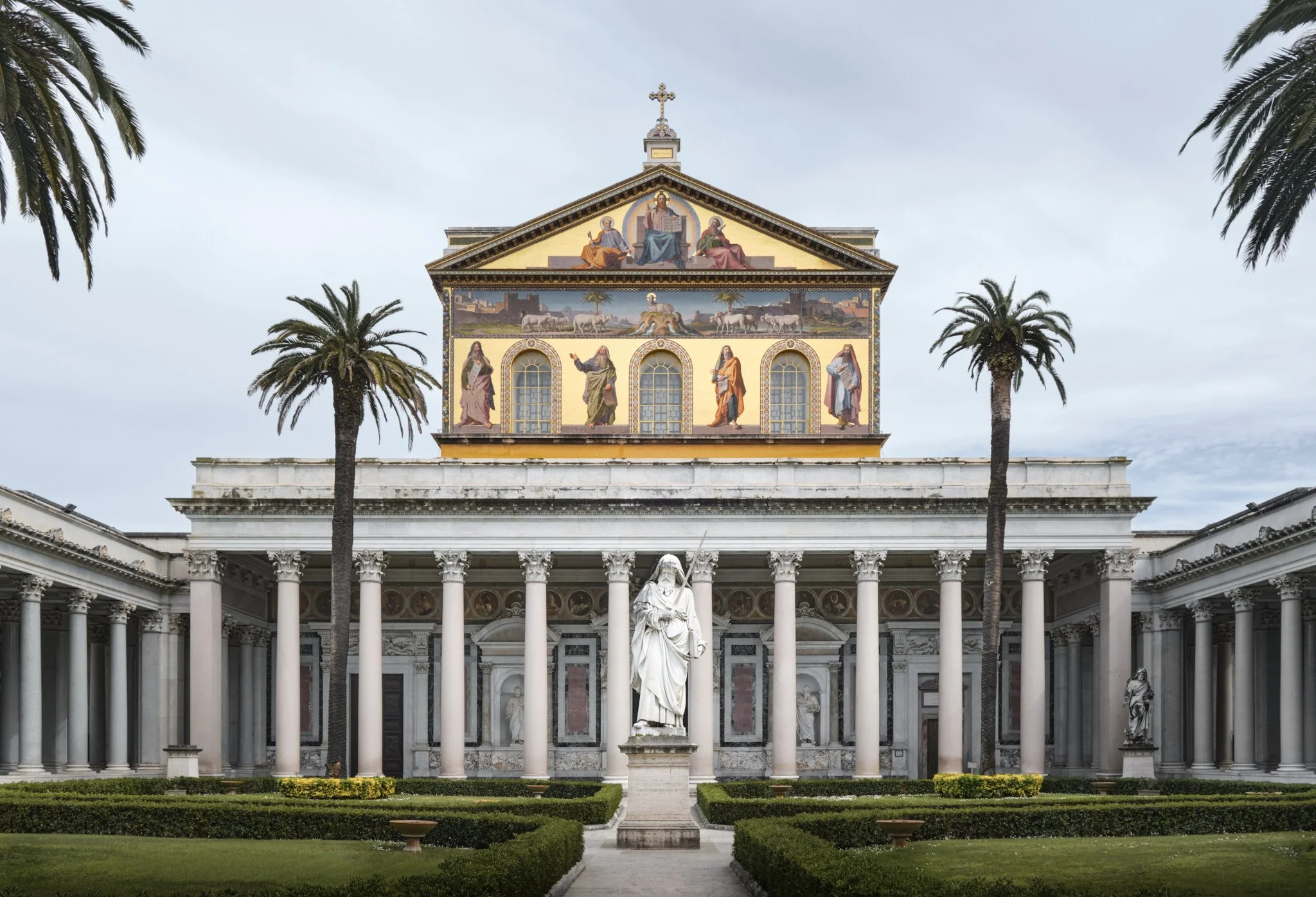 Full exterior facade of the Basilica di San Paolo Fuori le Mura with palm trees in the forecourt, the statue of Saint Paul, and the golden mosaic frieze above the entrance portico.
