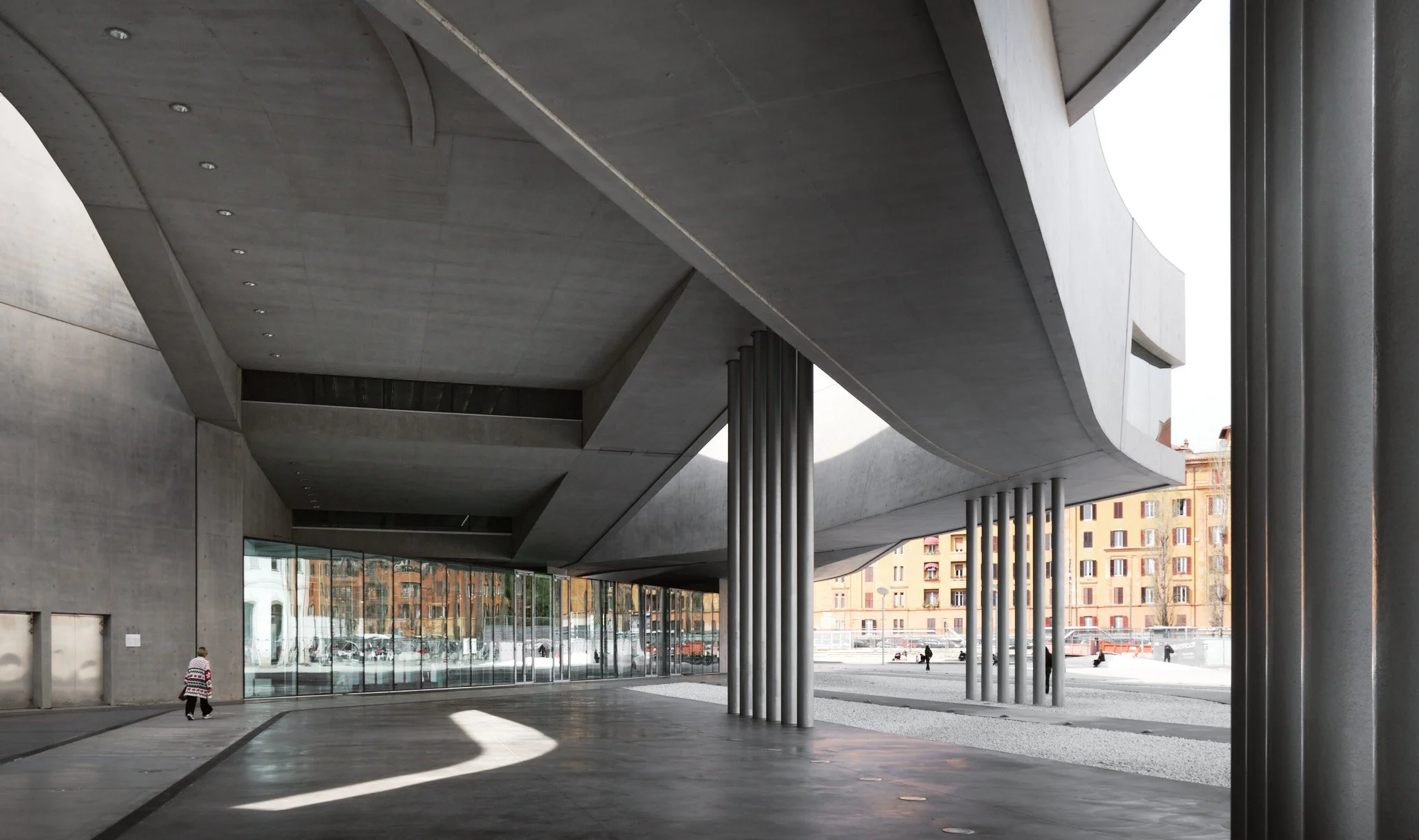Exterior covered gallery at MAXXI Rome with rhythmic concrete columns and a human figure providing scale, seen in natural light.