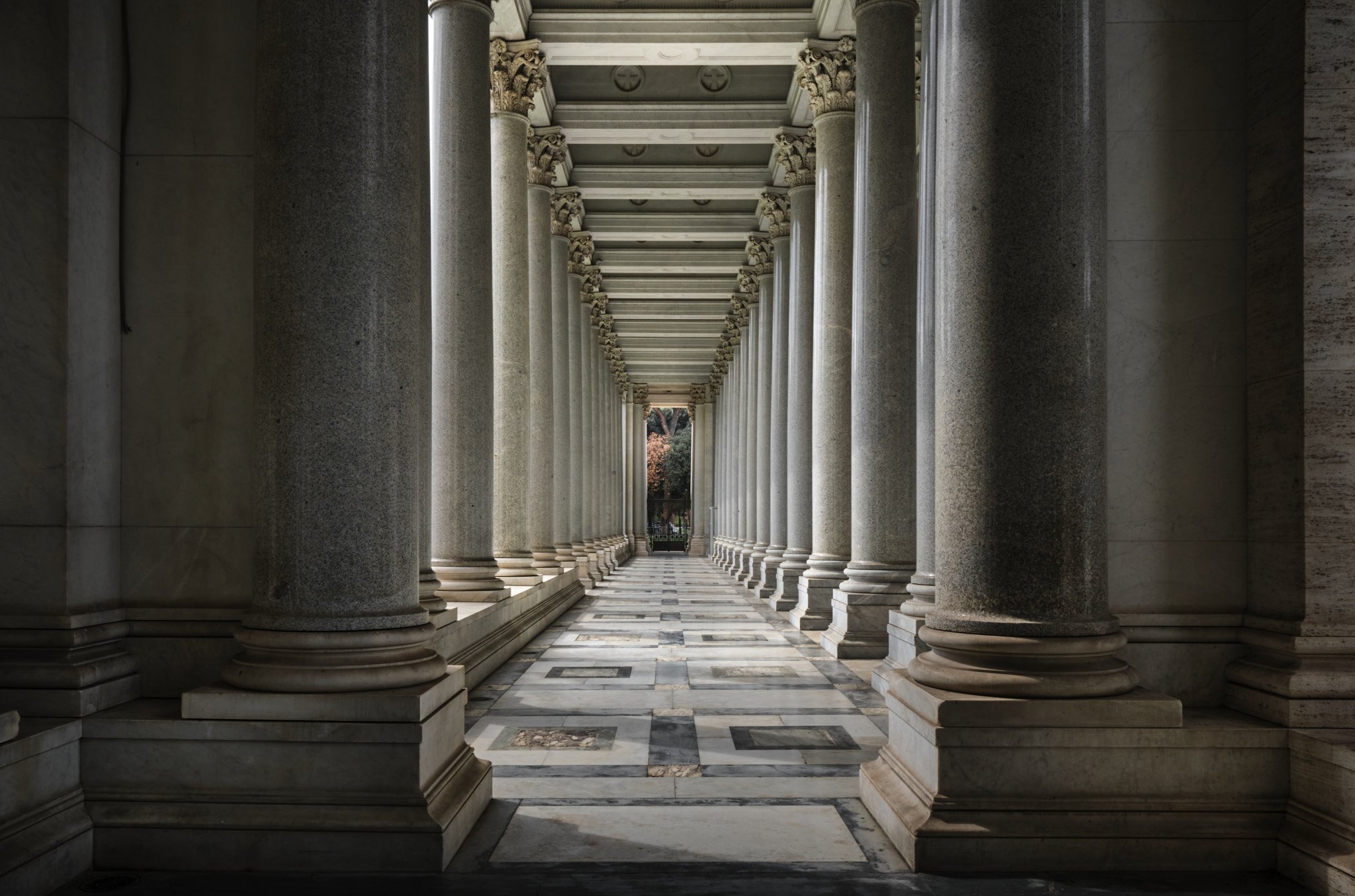 Long interior colonnade of the Basilica di San Paolo Fuori le Mura, dark granite columns receding toward a bright exit, small figure visible at the far end providing scale.