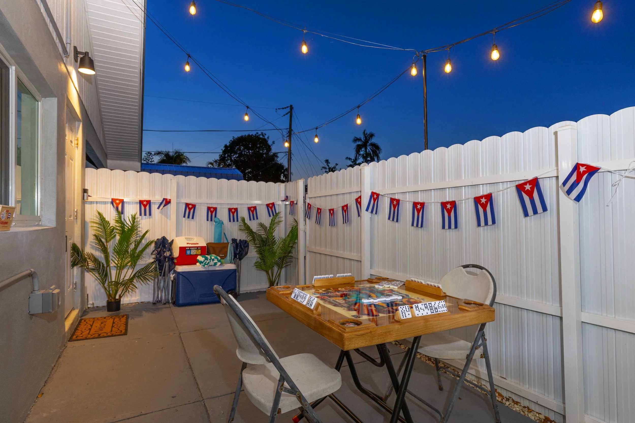 Backyard patio decorated for a celebration with Cuban flags, string lights, a table with dominoes, chairs, and potted plants.